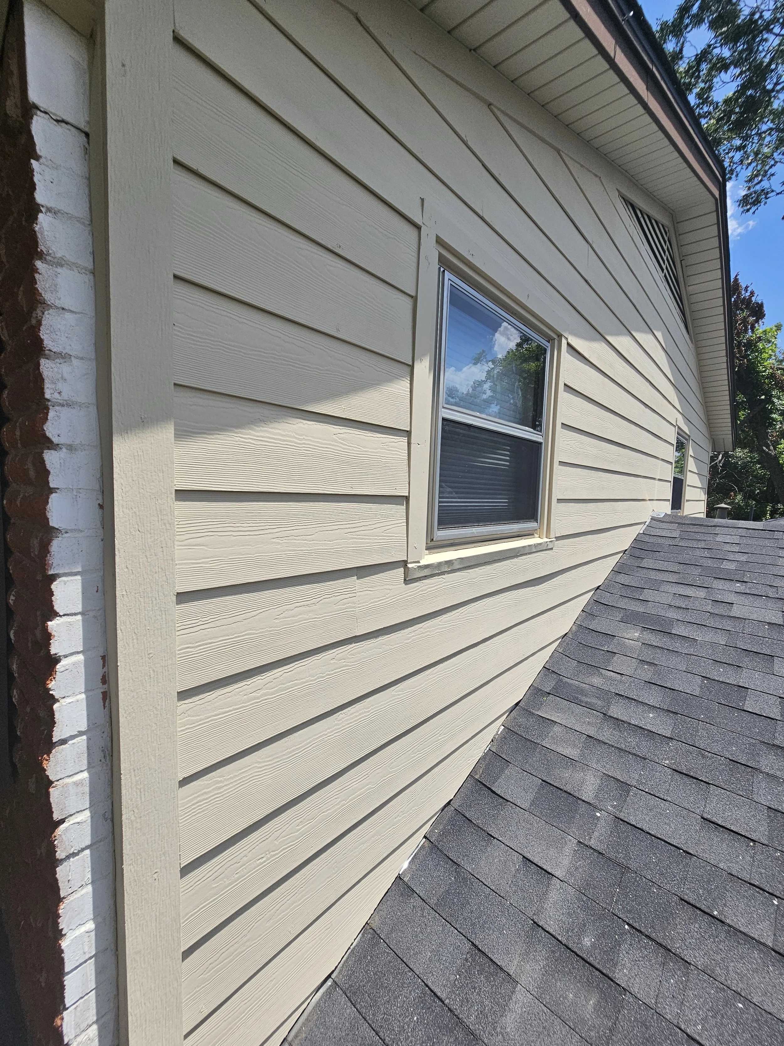 Exterior view of a house with beige horizontal siding, double window, and black shingle roof. Part of a brick chimney is visible on the left side, with trees and a blue sky in the background.