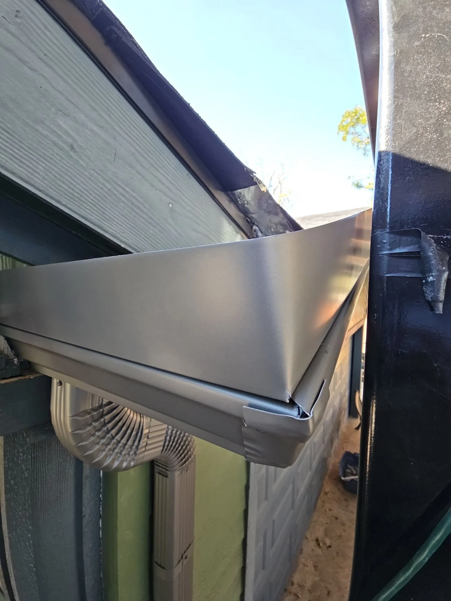 A stainless steel outdoor air conditioning unit on a house exterior wall, with a large white plastic air intake and ducts below it.