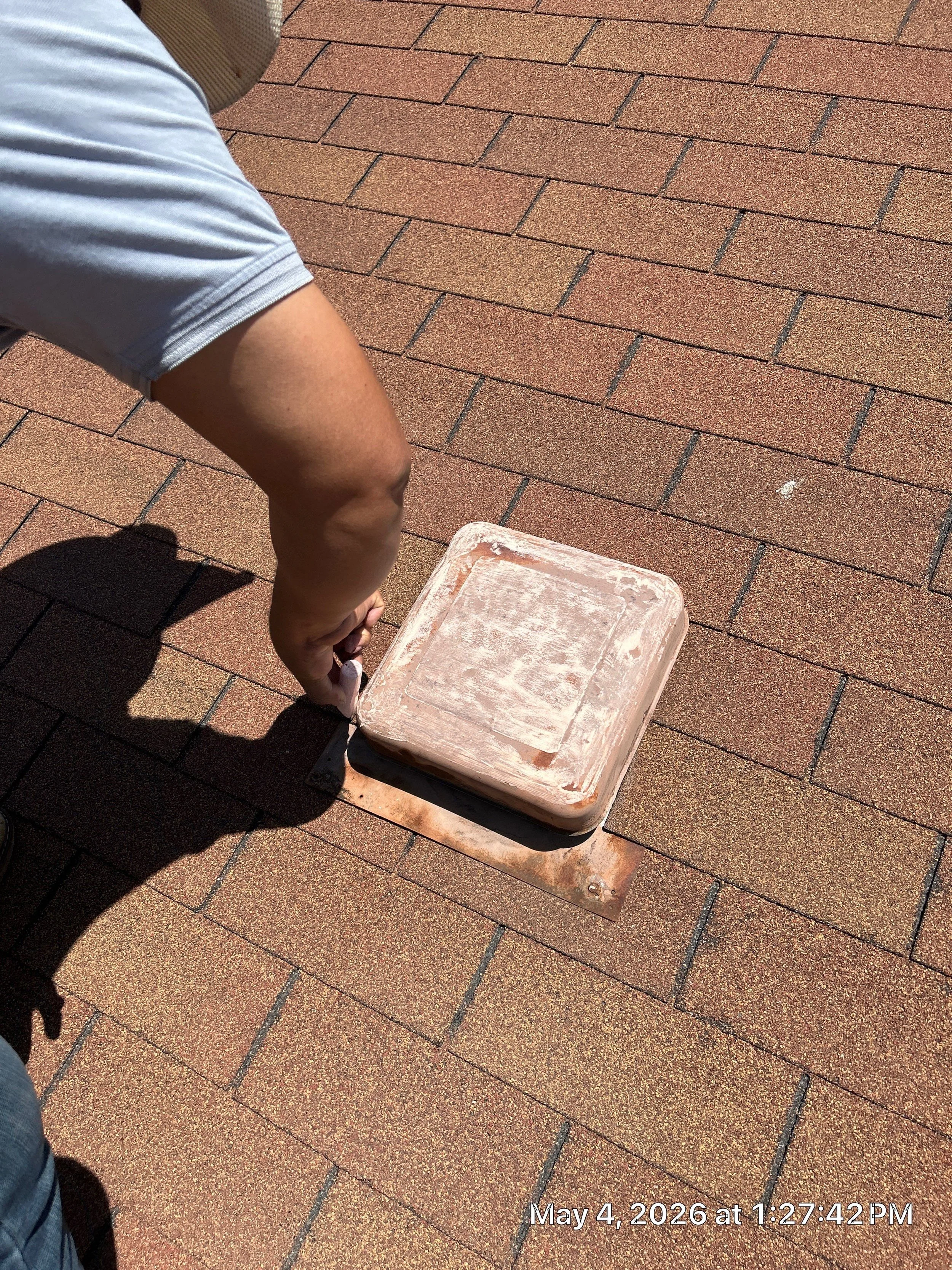 A person wearing a white T-shirt and denim shorts is pointing at a rusty metal vent on a brick sidewalk or roof. The date and time in the bottom right corner show May 4, 2026, at 1:27:42 PM.