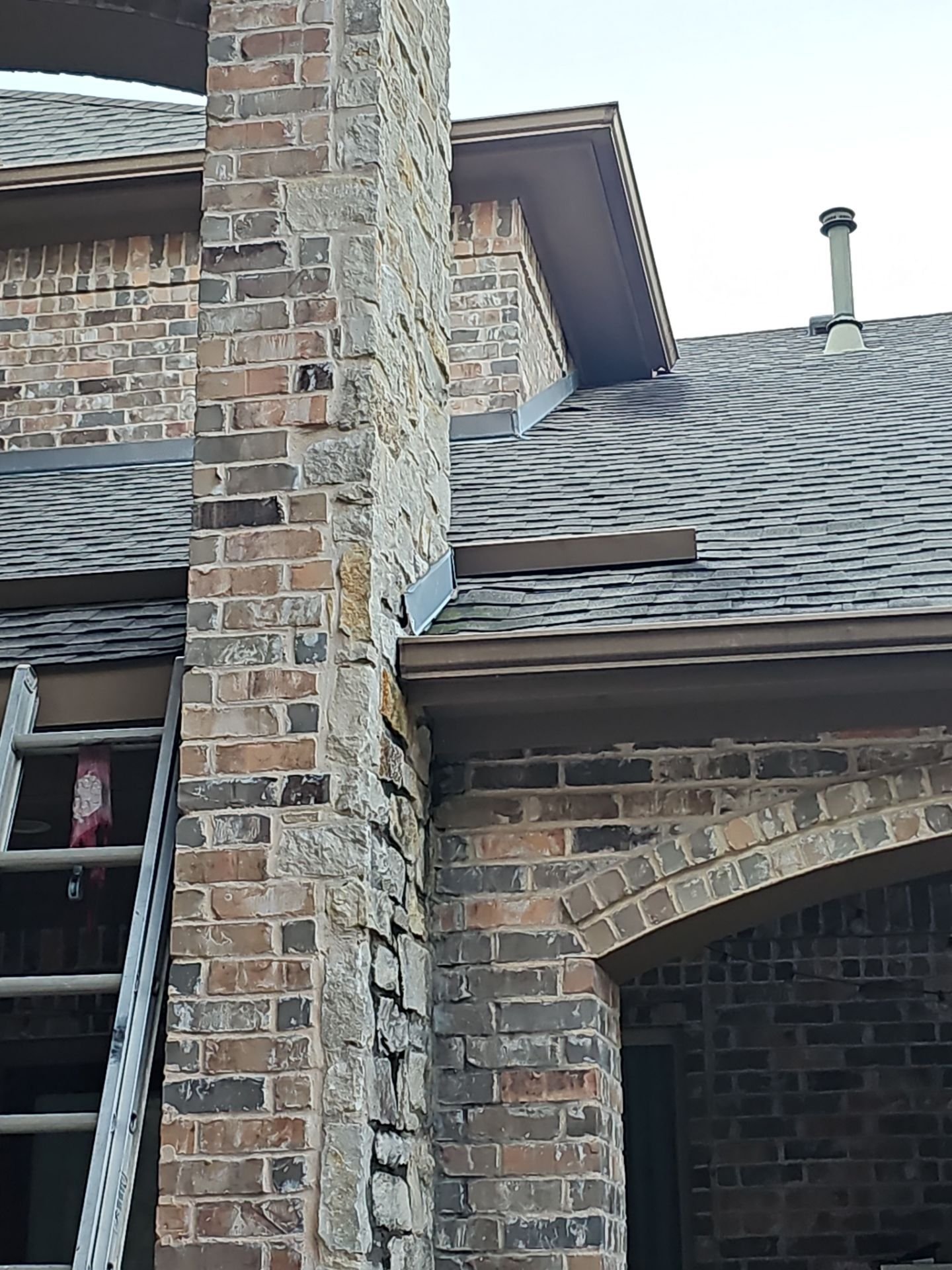 Close-up view of a brick house's exterior wall, showing a stone chimney, a window with a ladder leaning against it, and part of the roof with shingles and a vent pipe.