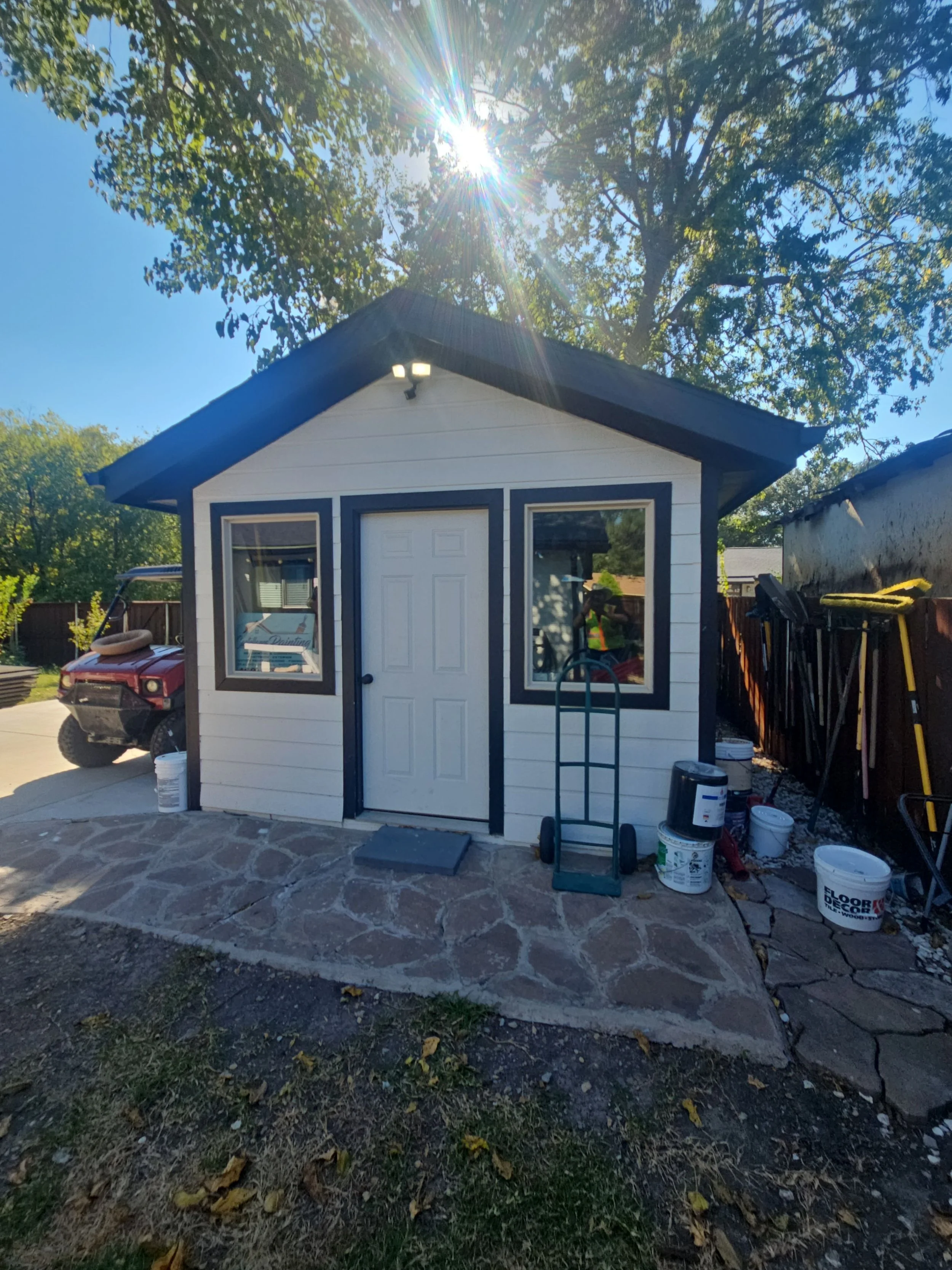 Backyard shed with a white door and two windows, construction supplies and tools surrounding it, and trees with sunlight overhead.
