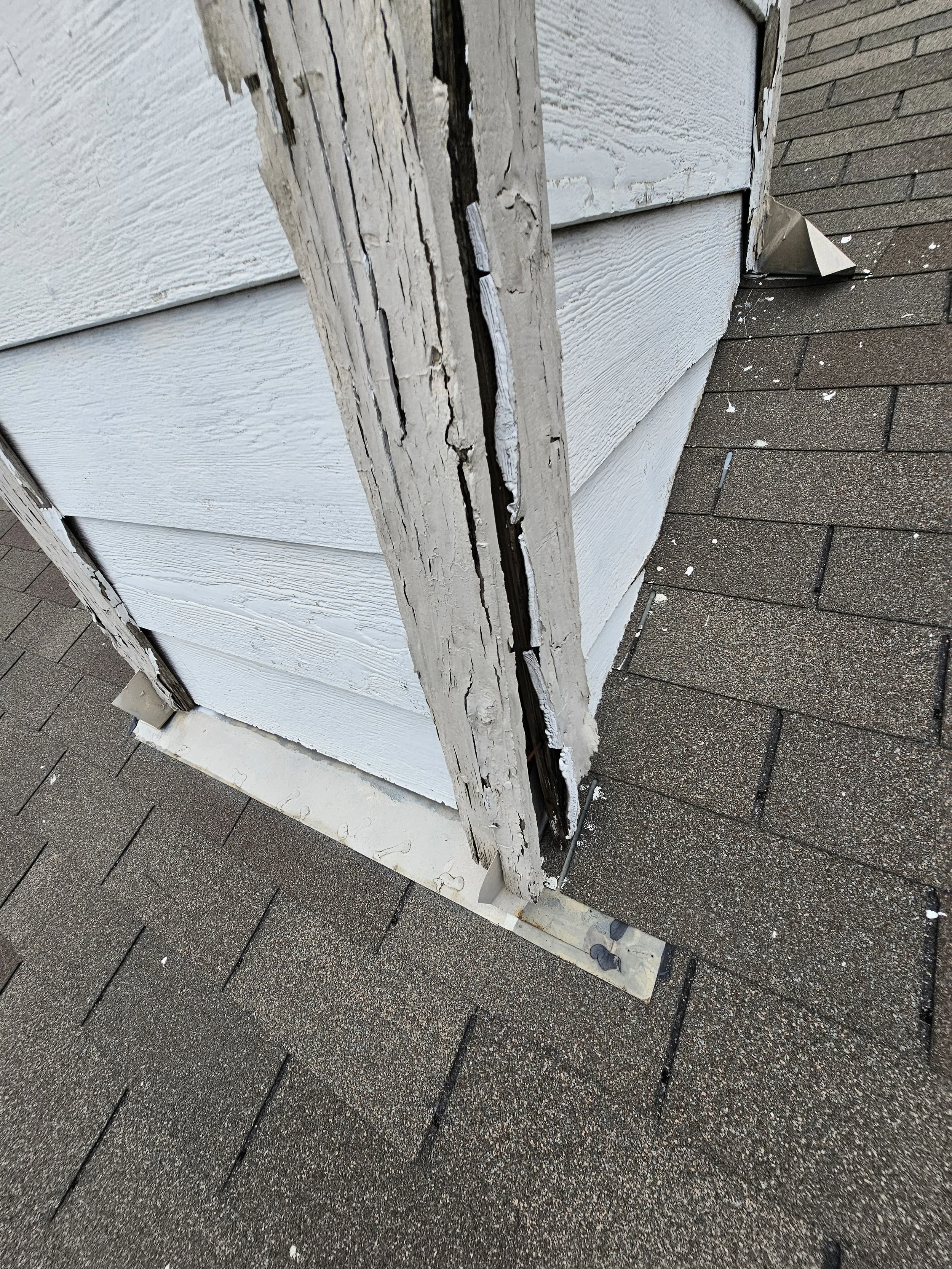 Close-up of a damaged section of a house exterior with chipped and peeling white siding, exposing the underlying wood that appears rotten and deteriorated, alongside a brown shingled roof.