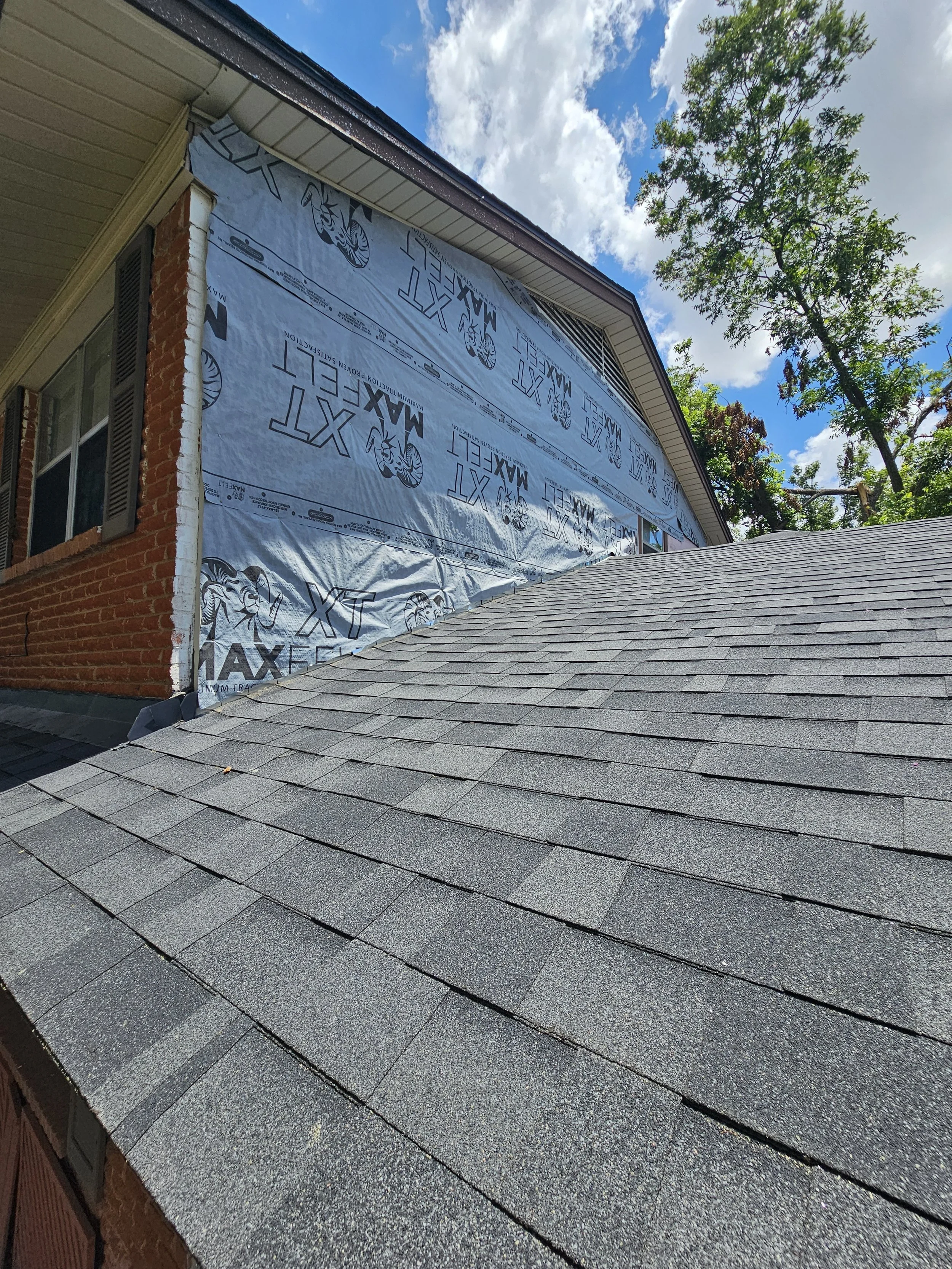 The image shows a house with part of the exterior wrapped in weather-resistant house wrap featuring branding and logos. The house has a brick exterior, window shutters, and asphalt shingle roofing. The sky is partly cloudy with white clouds and a blu