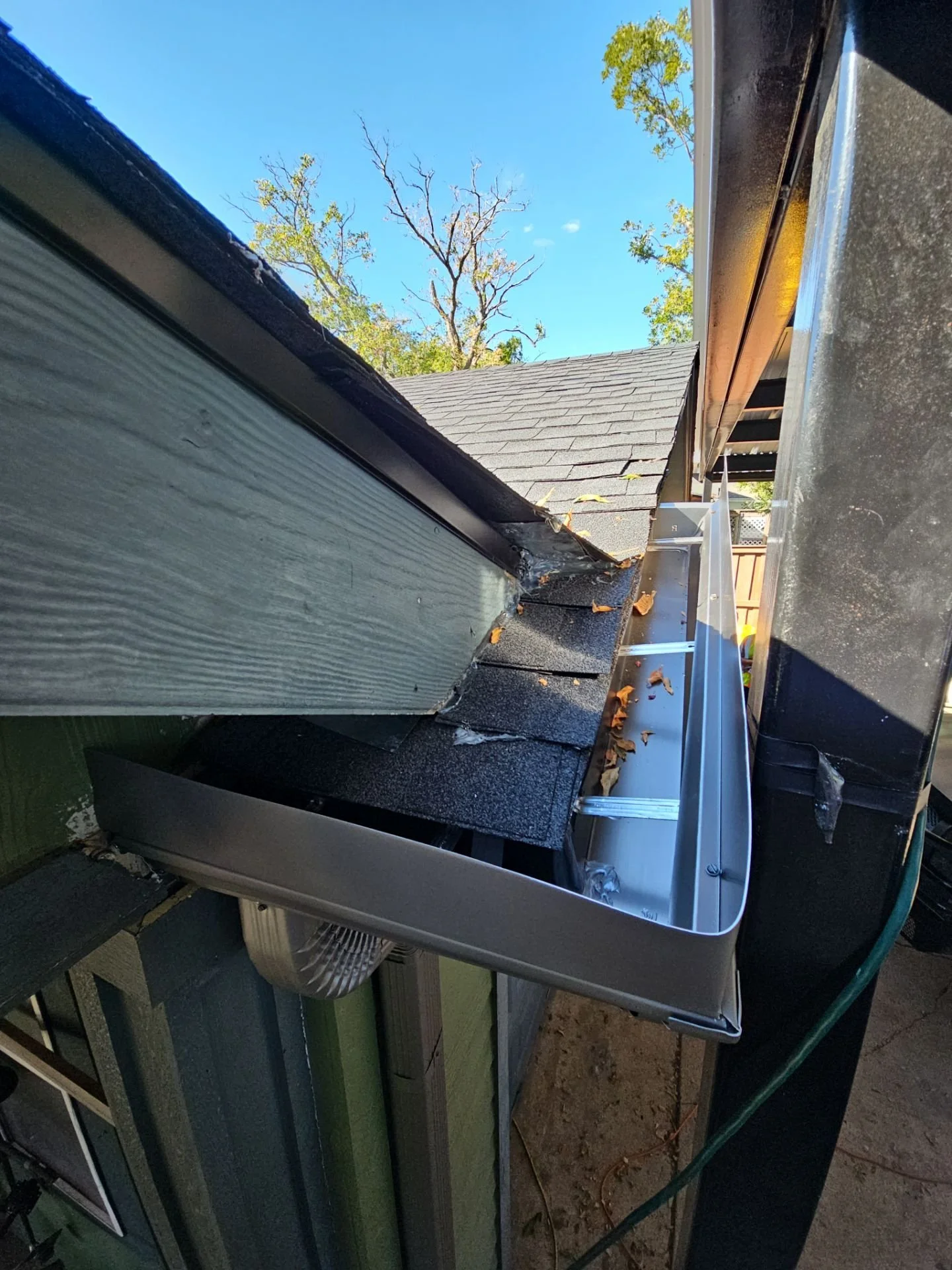 View of a roof with a gutter, shingles, and a ladder, with trees and a clear blue sky in the background.