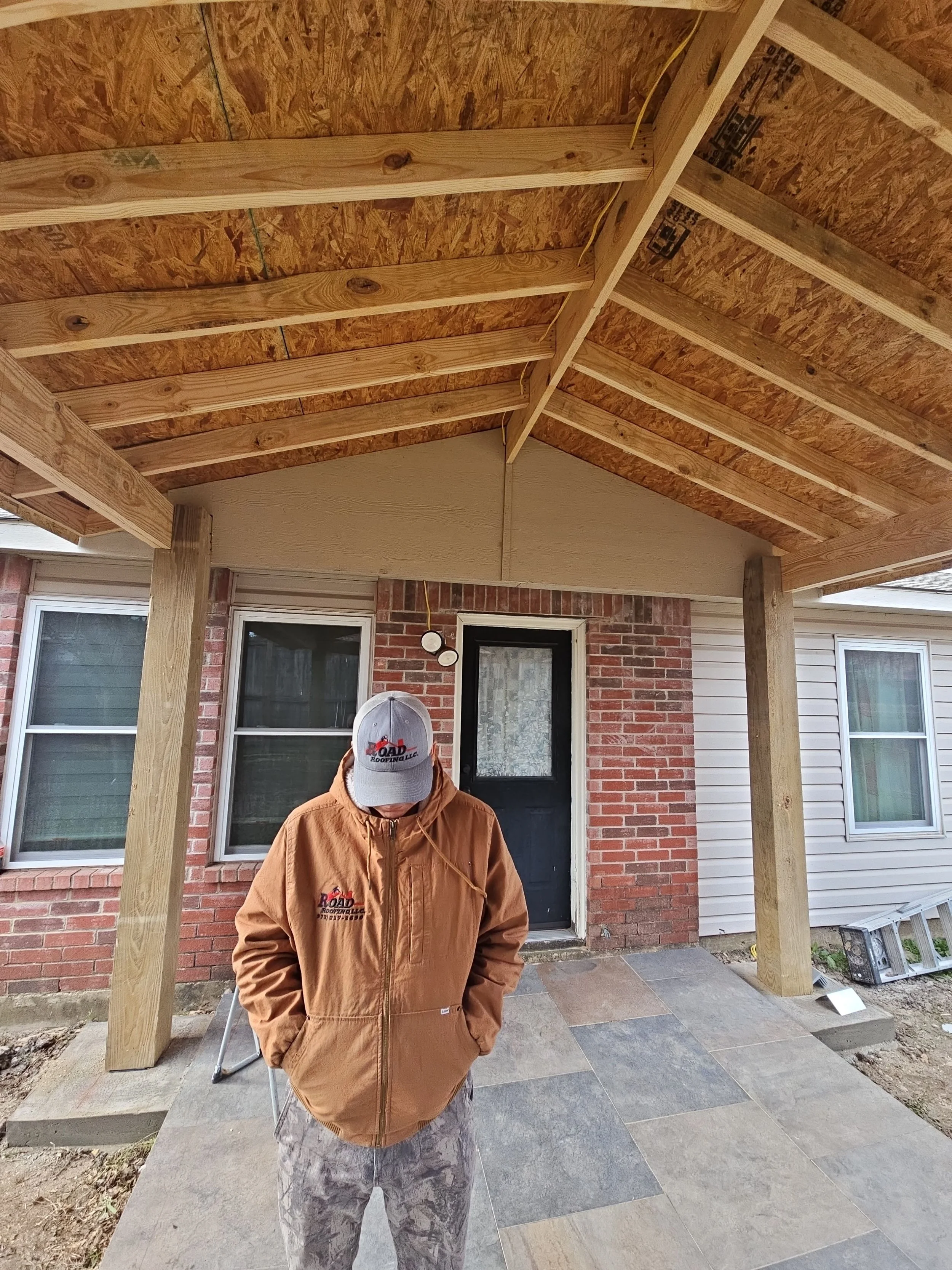 A man wearing a brown jacket and camouflage pants standing outside under construction or renovation. He is wearing a gray cap with a logo and is looking downward with his face partially obscured. The background shows a brick house with windows and a 