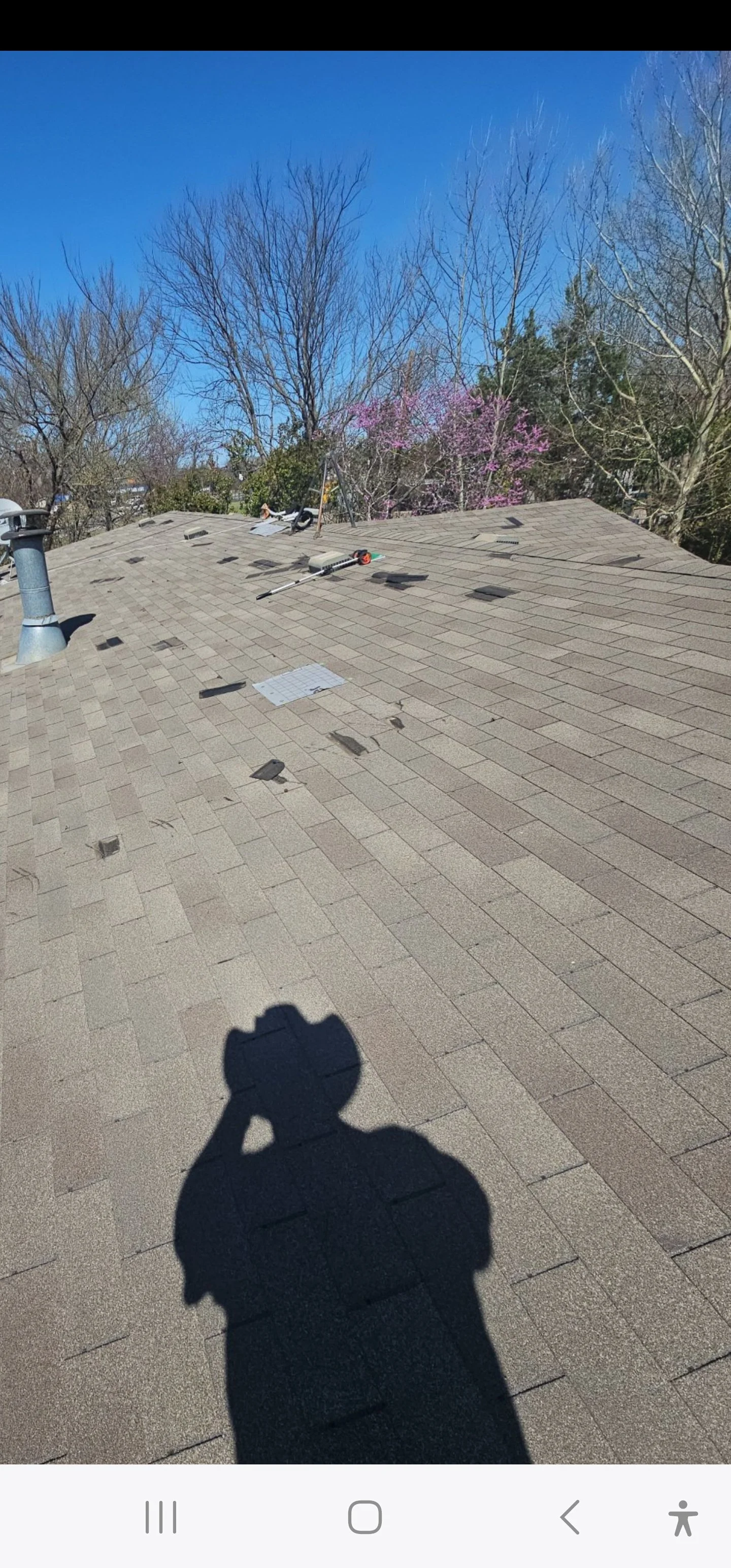View of a residential rooftop with vents, tools, and patches of missing shingles, under a clear blue sky with leafless trees in the background.