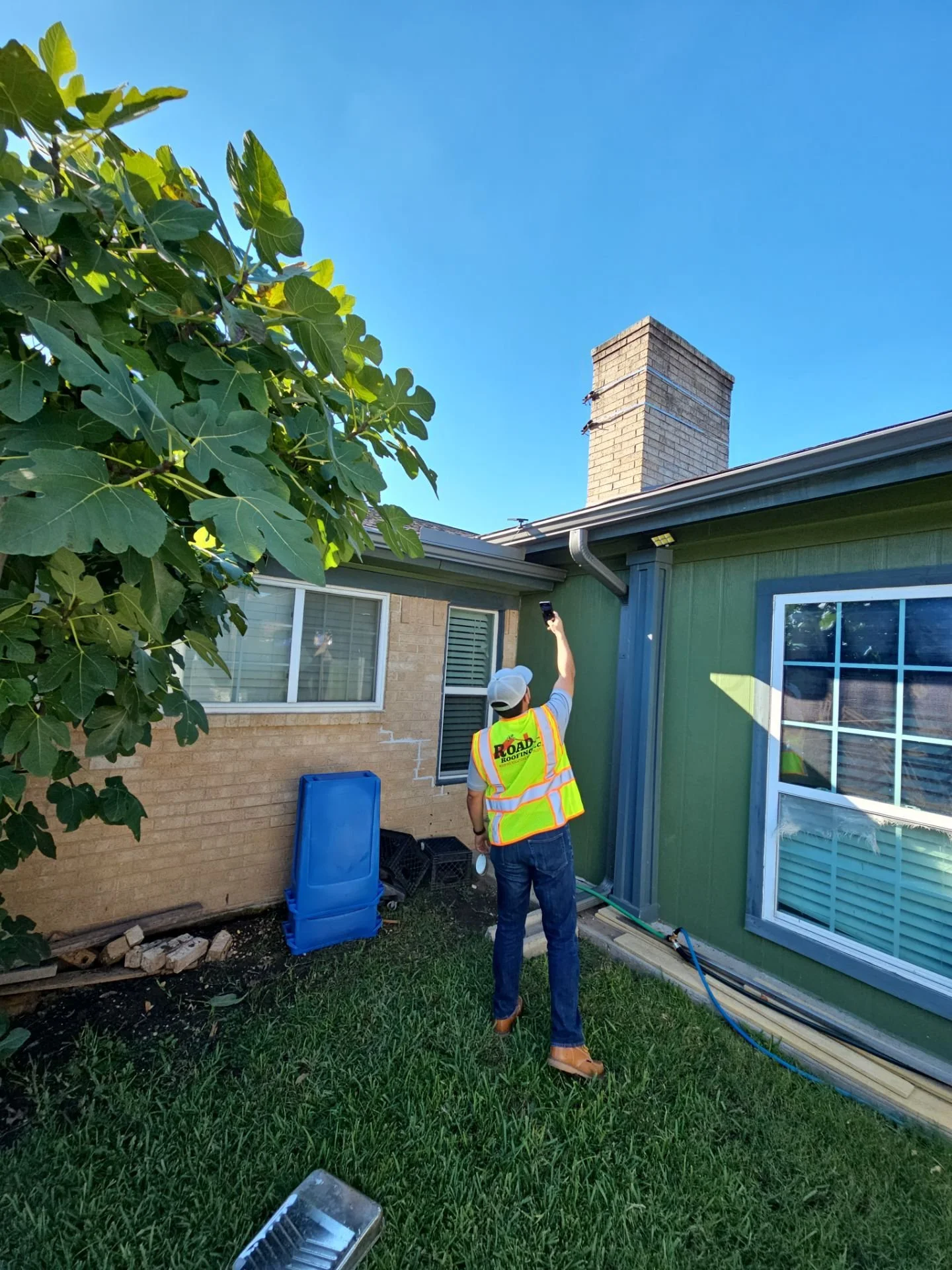 A construction worker in a yellow high-visibility vest and white cap is standing outside a house, pointing a black device towards the chimney on the roof. The house has green siding, a large window with blinds, and a brick chimney. There are some con