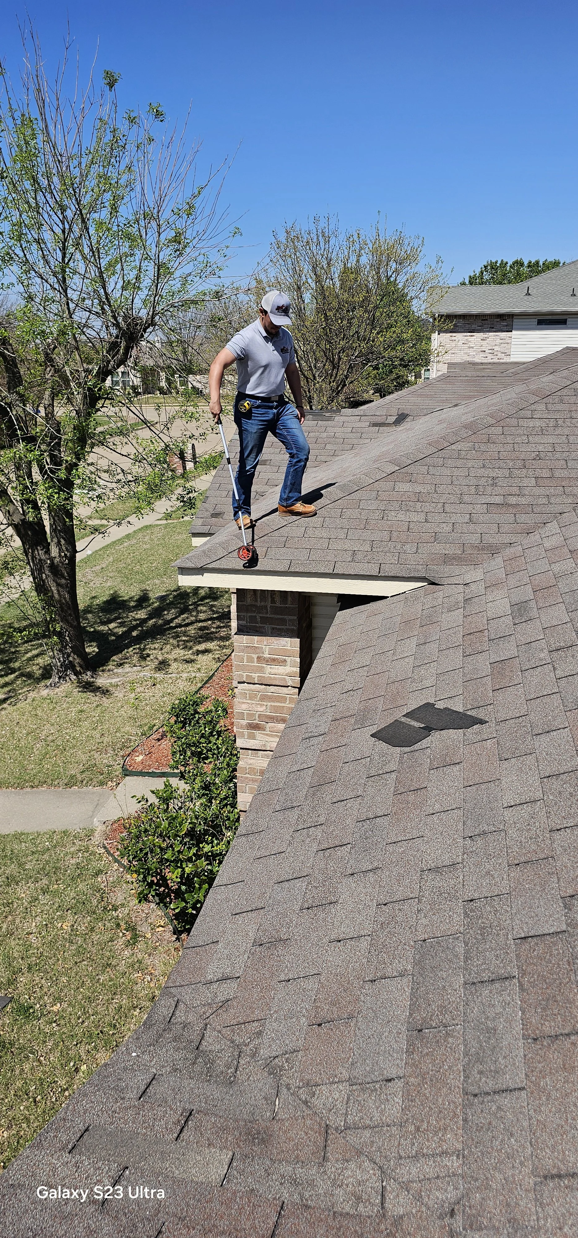 A man standing on the edge of a house roof, inspecting or working on the shingles, with trees and neighboring houses in the background on a sunny day.