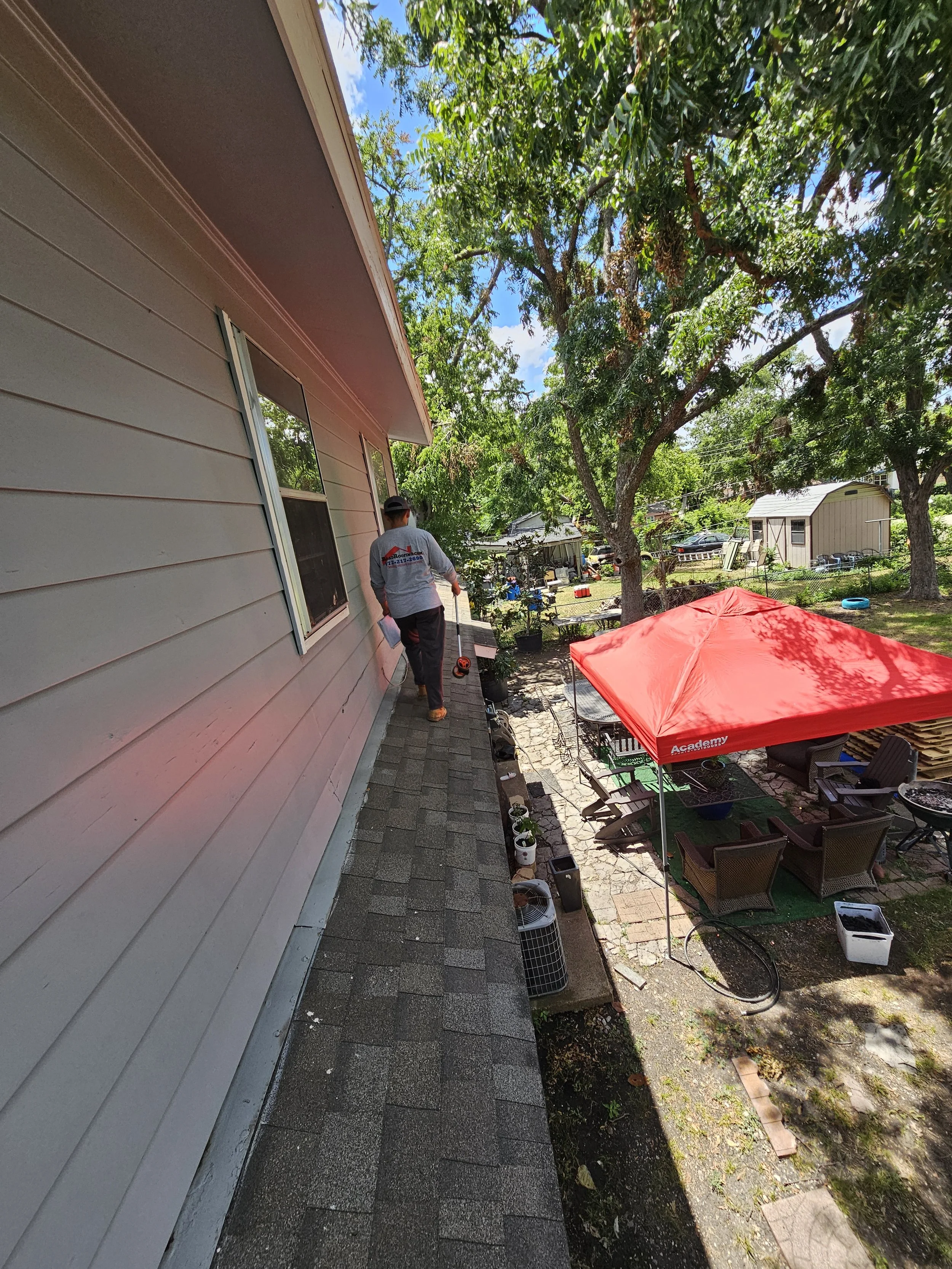 A man with a gray shirt walking on a brick porch alongside a house with beige siding and two windows. The yard has trees, outdoor furniture, a red canopy, and various items scattered around on a sunny day.
