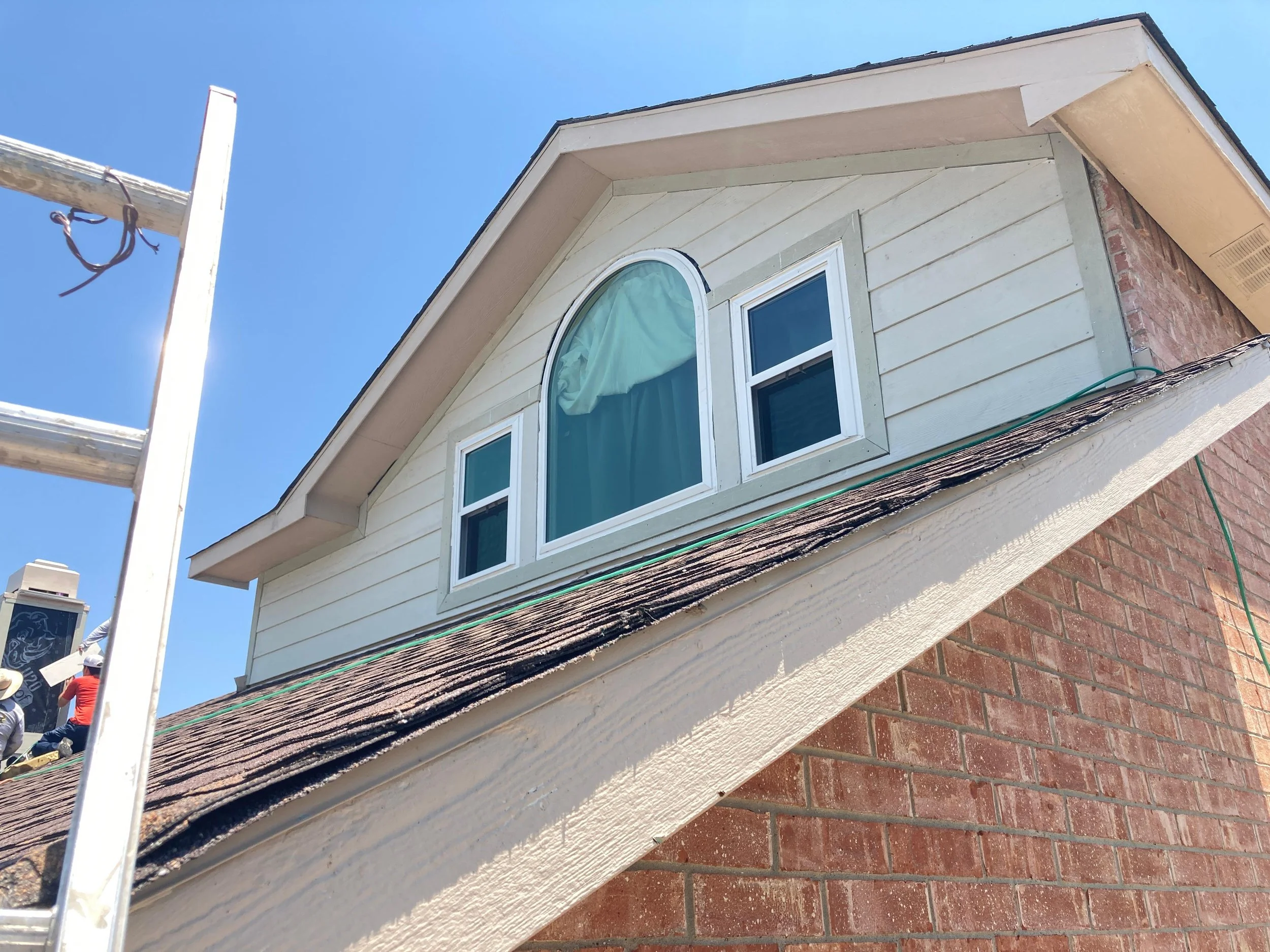 The image shows the upper part of a house with beige siding, an arched window, and a brick lower wall. A ladder is positioned against the house, and a person in a red shirt and white cap is visible at the bottom left corner, likely working on the ext