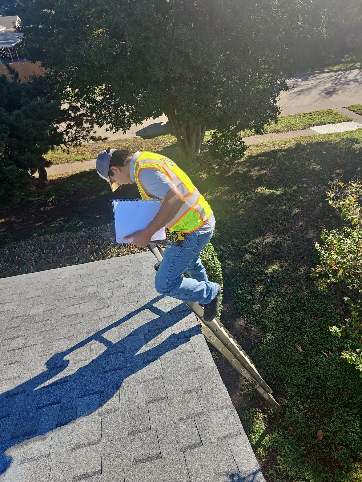 A man in a high-visibility safety vest, gray hat, and jeans is standing on a ladder on a residential sidewalk, writing or taking notes on a clipboard. He is shadowed on a paved path, with trees and shrubs nearby.