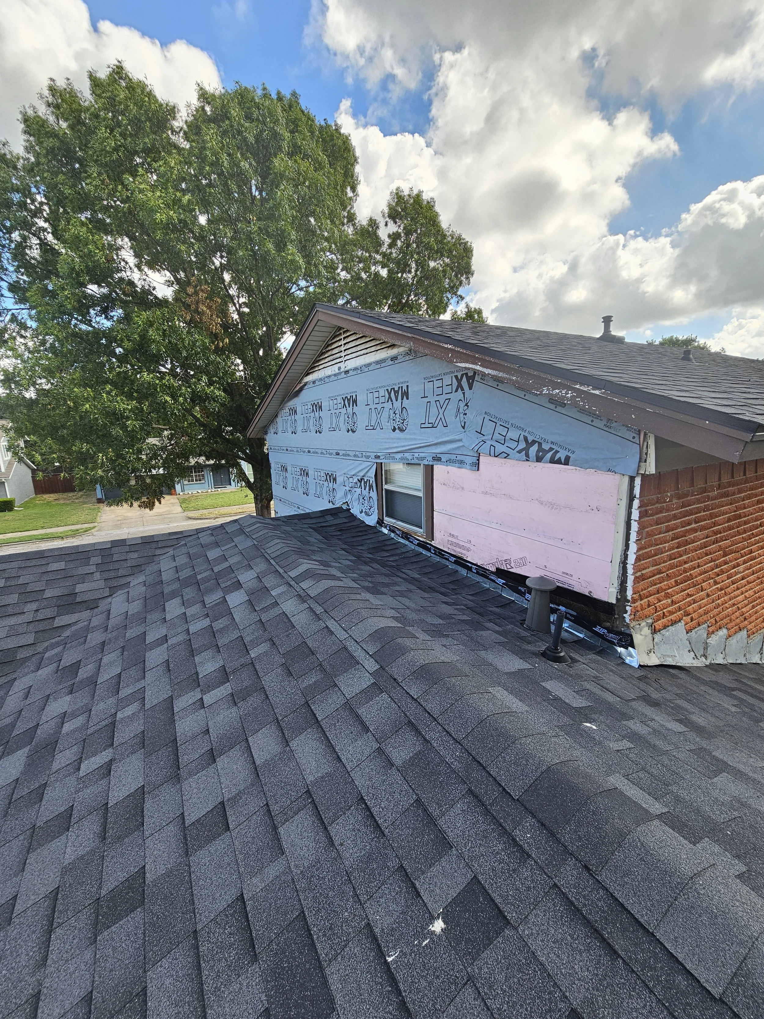 A house with a partially work-in-progress exterior, with a roof, a window, and siding under construction, on a street with large trees and a blue sky with clouds.