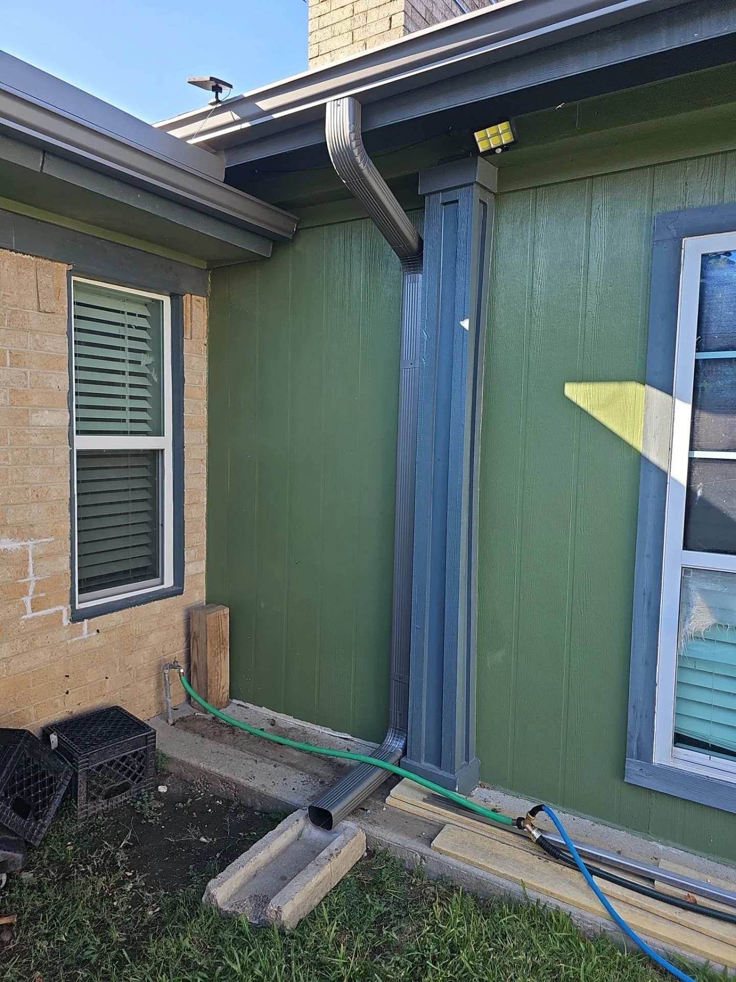 The image shows the exterior corner of a building with green and brick walls, a window with blinds, and construction materials including wooden blocks, a green garden hose, a black crate, and some black tubing on the ground, suggesting ongoing constr