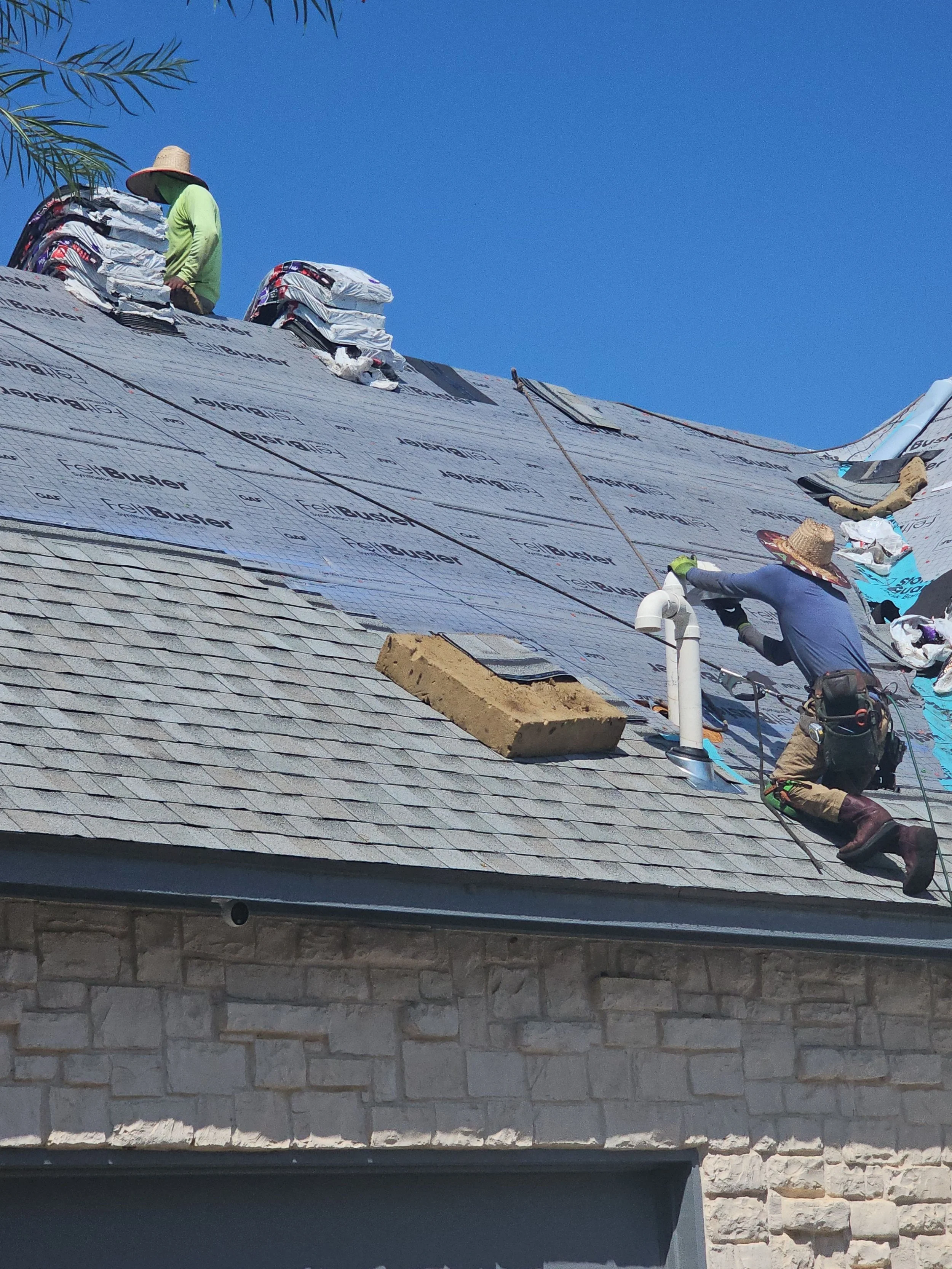 Roofing workers installing shingles on a sloped roof during the daytime, with one worker in a hat and another in a green shirt.