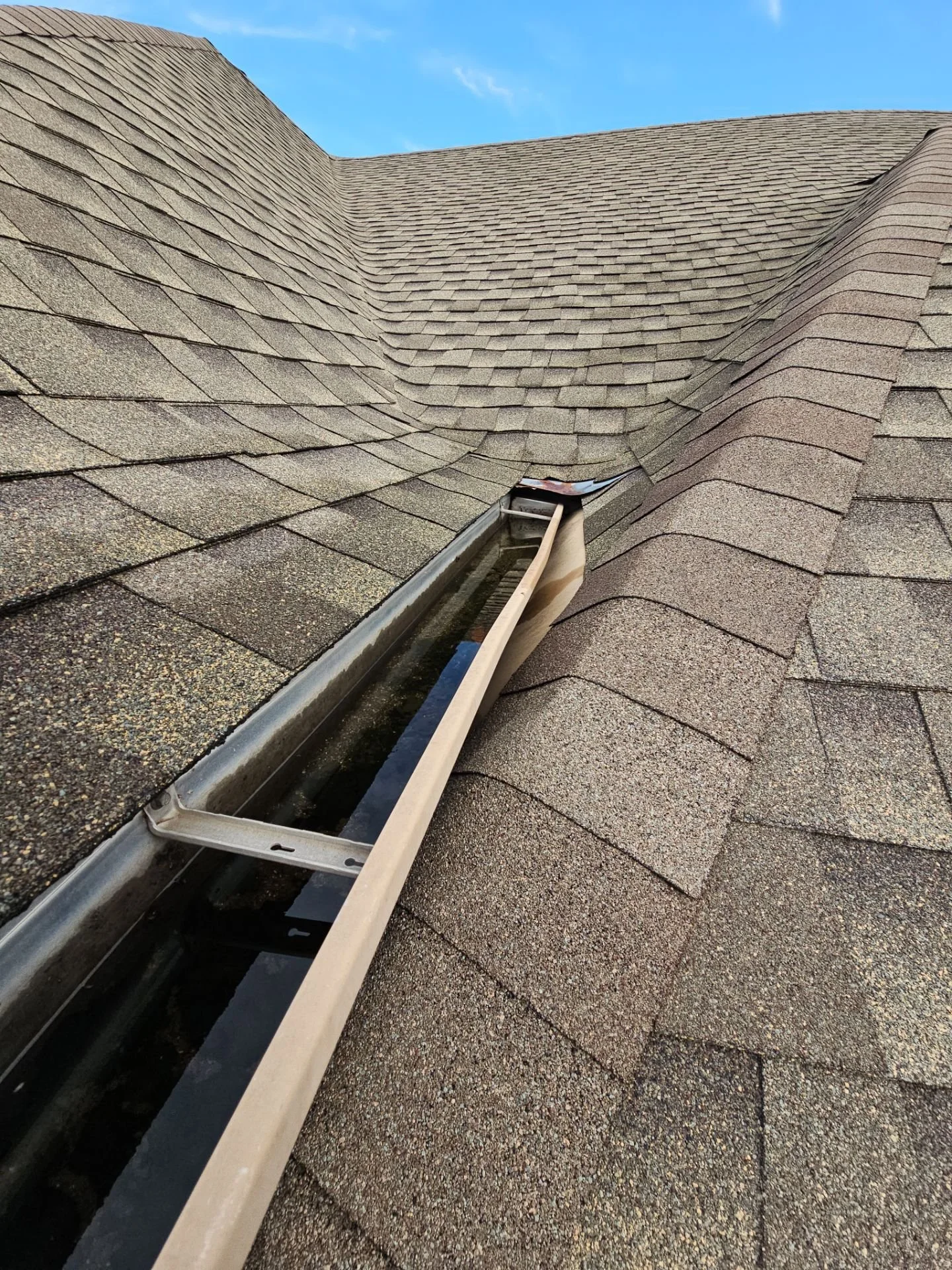 View of a shingled roof with a damaged section near the gutter, where a skylight or vent cover is missing.