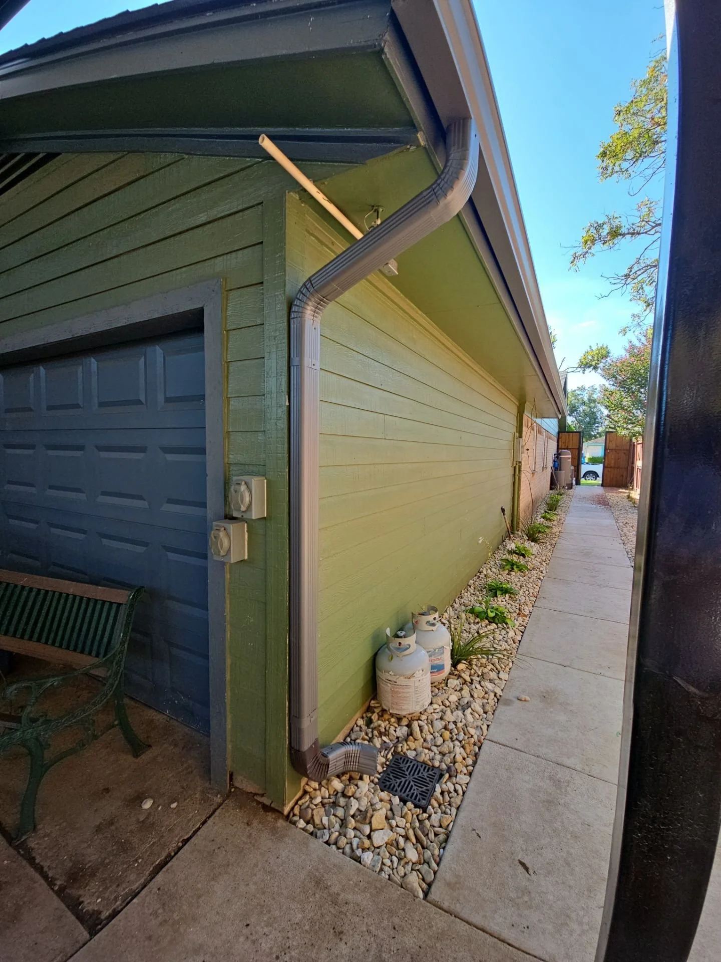 Side view of a green house with a garage door, rain gutter system, and propane tanks outside on a small gravel area, with a sidewalk running alongside and trees in the background under a blue sky.