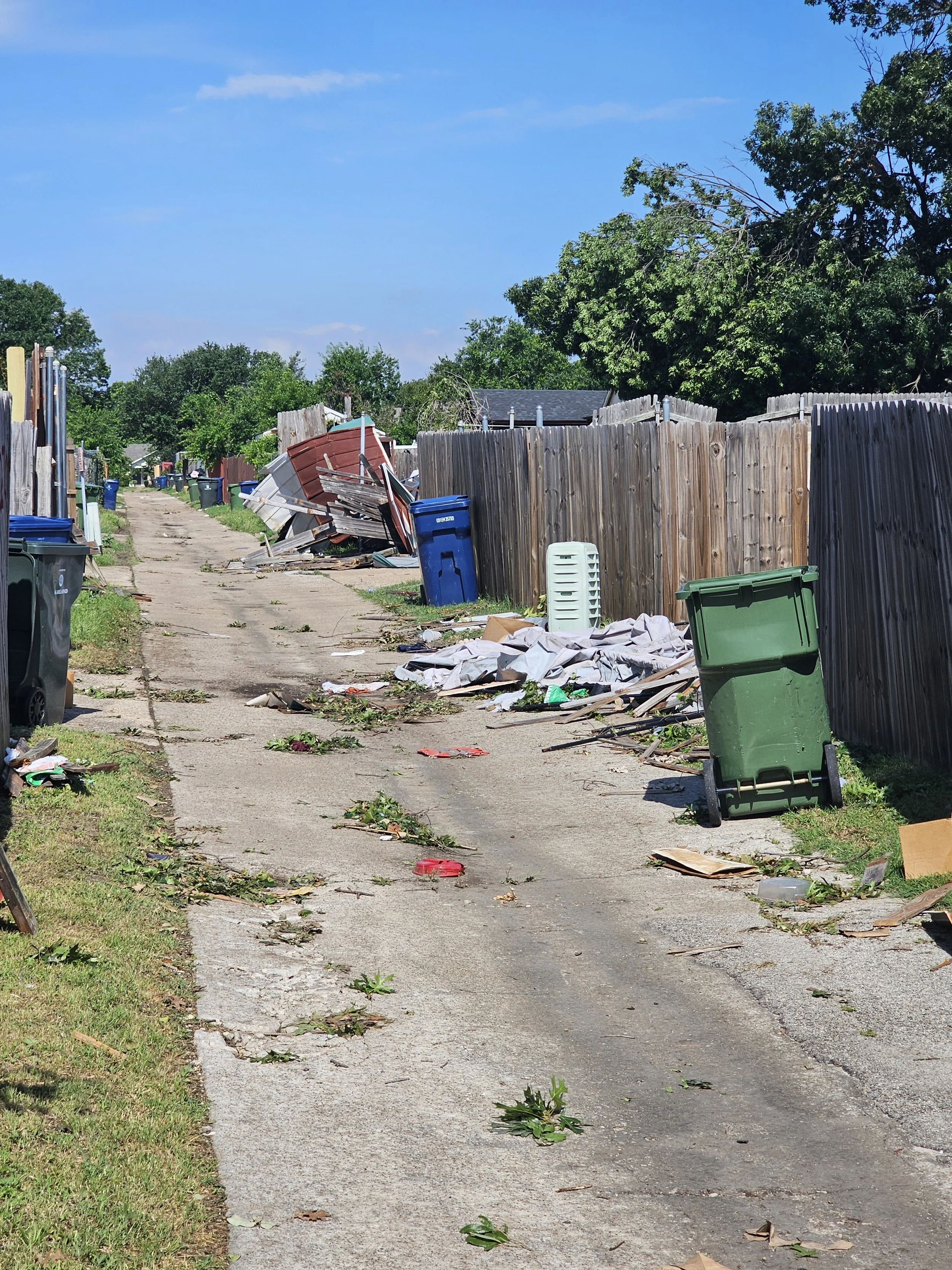 A residential alleyway with trash and debris scattered along the sidewalk and yard, a wooden fence on the right, and trash cans, some knocked over, on both sides of the walkway. Trees and houses are visible in the background under a clear blue sky.