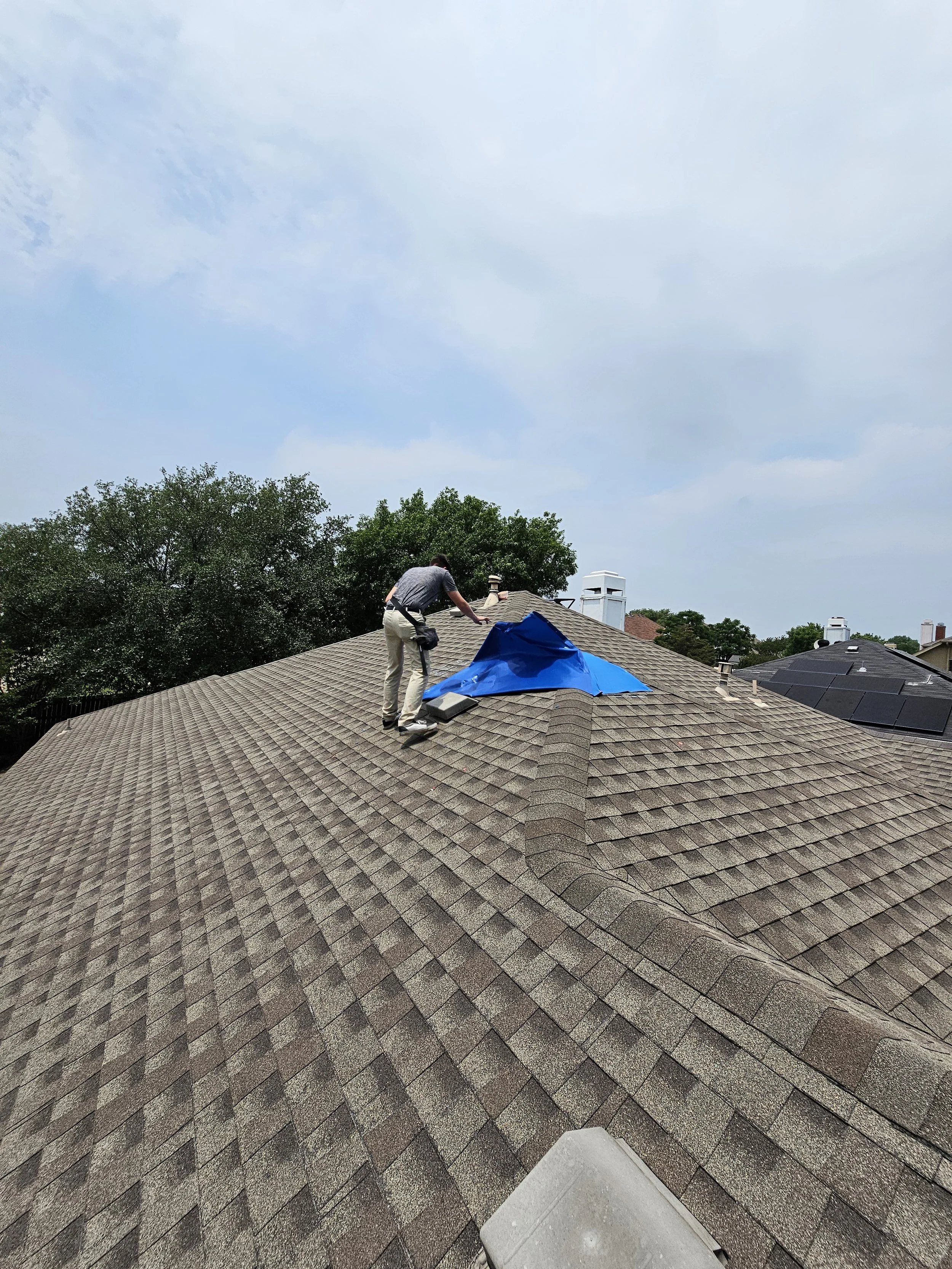 A person on a roof repairing or inspecting the shingles, with a blue tarp nearby, under a partly cloudy sky with trees in the background.
