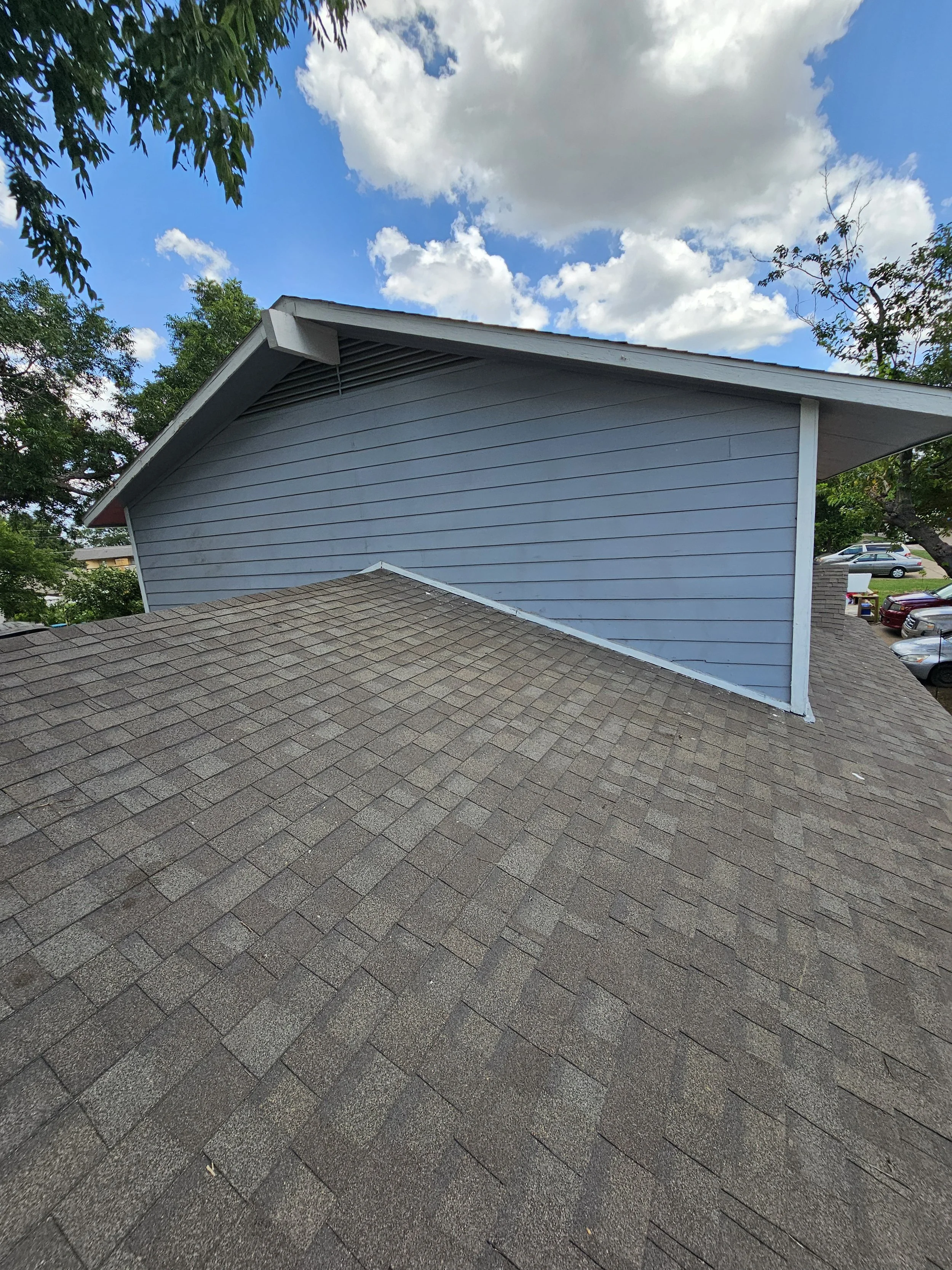 View of a rooftop with gray asphalt shingles, a side of a house with light blue siding, and a neighboring parking lot with several cars. Light clouds are visible in a partly cloudy sky, with some trees around.