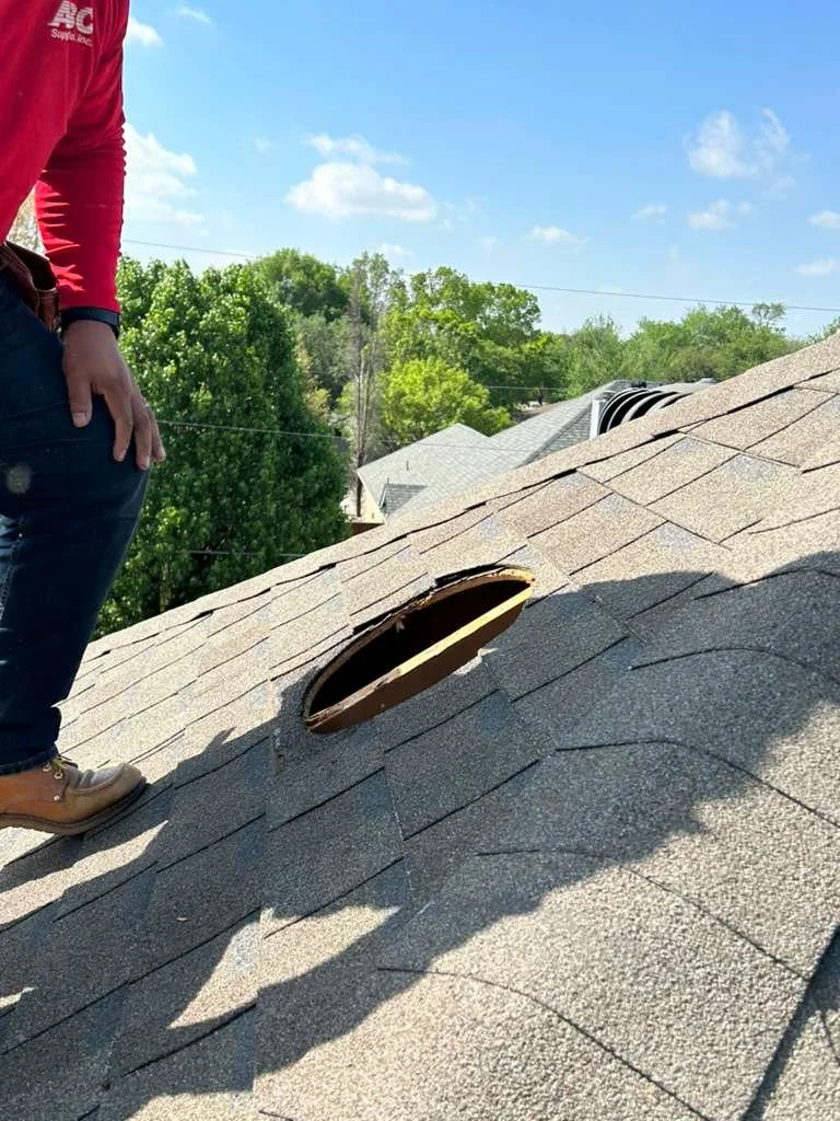 Person standing on a residential roof with shingles, near a circular hole in the roof, with trees and houses in the background.