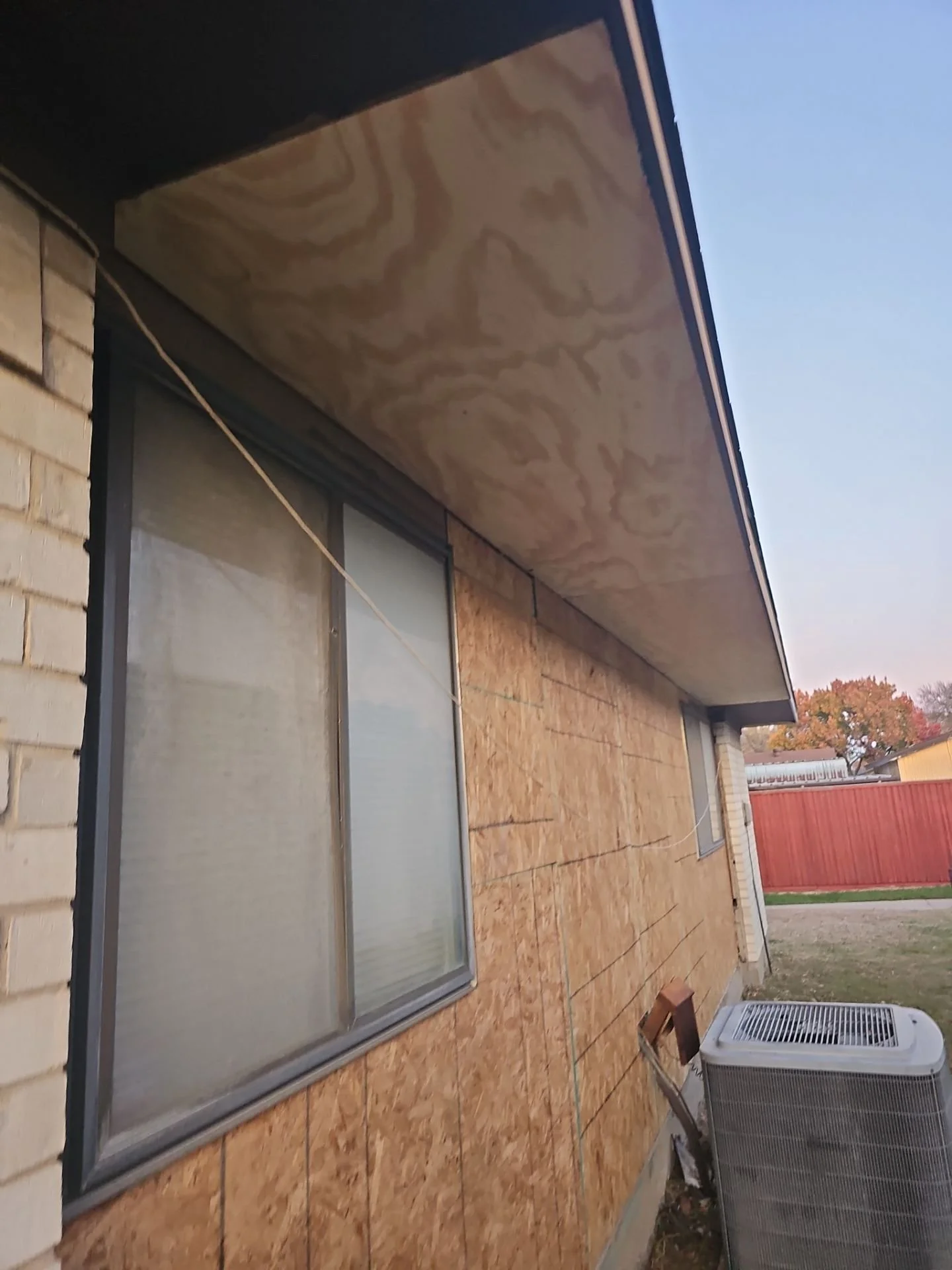 Side of a house with a brick wall, large sliding glass door, plywood paneling on the wall, and an air conditioning unit outside, with a red wooden fence and trees in the background at sunset.
