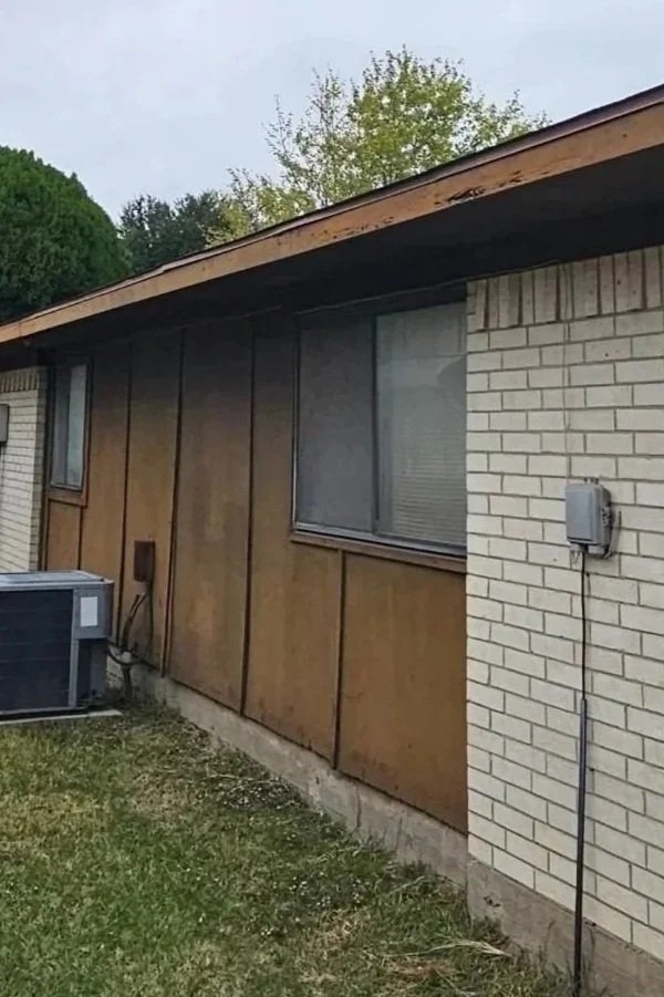 Side view of a house with wood paneling and white brick wall, two windows, an outdoor unit, and electrical fixtures with a grassy yard.