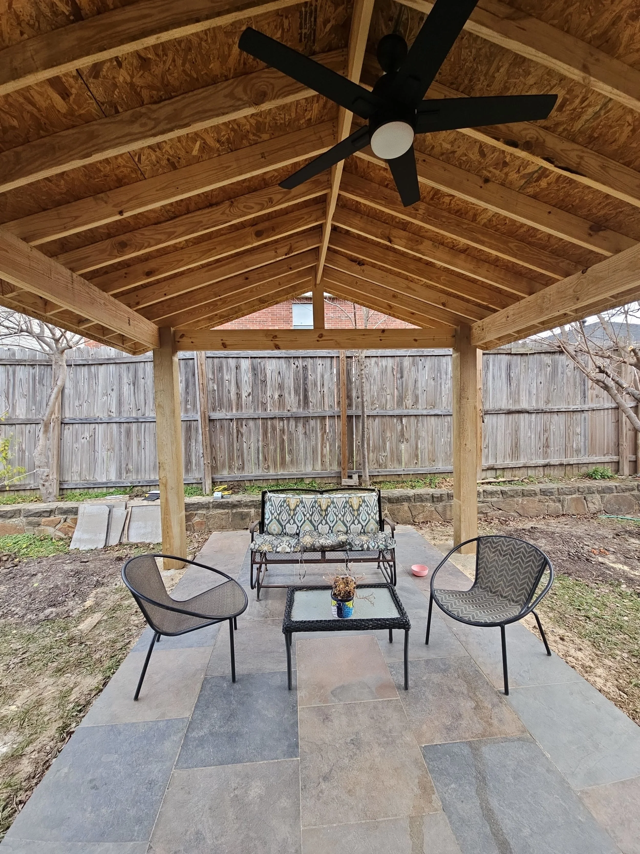 Backyard patio with four black chairs and a small black table with a potted plant, covered by a wooden pergola with a ceiling fan, wooden fence in the background, and trees and brick house visible beyond the fence.