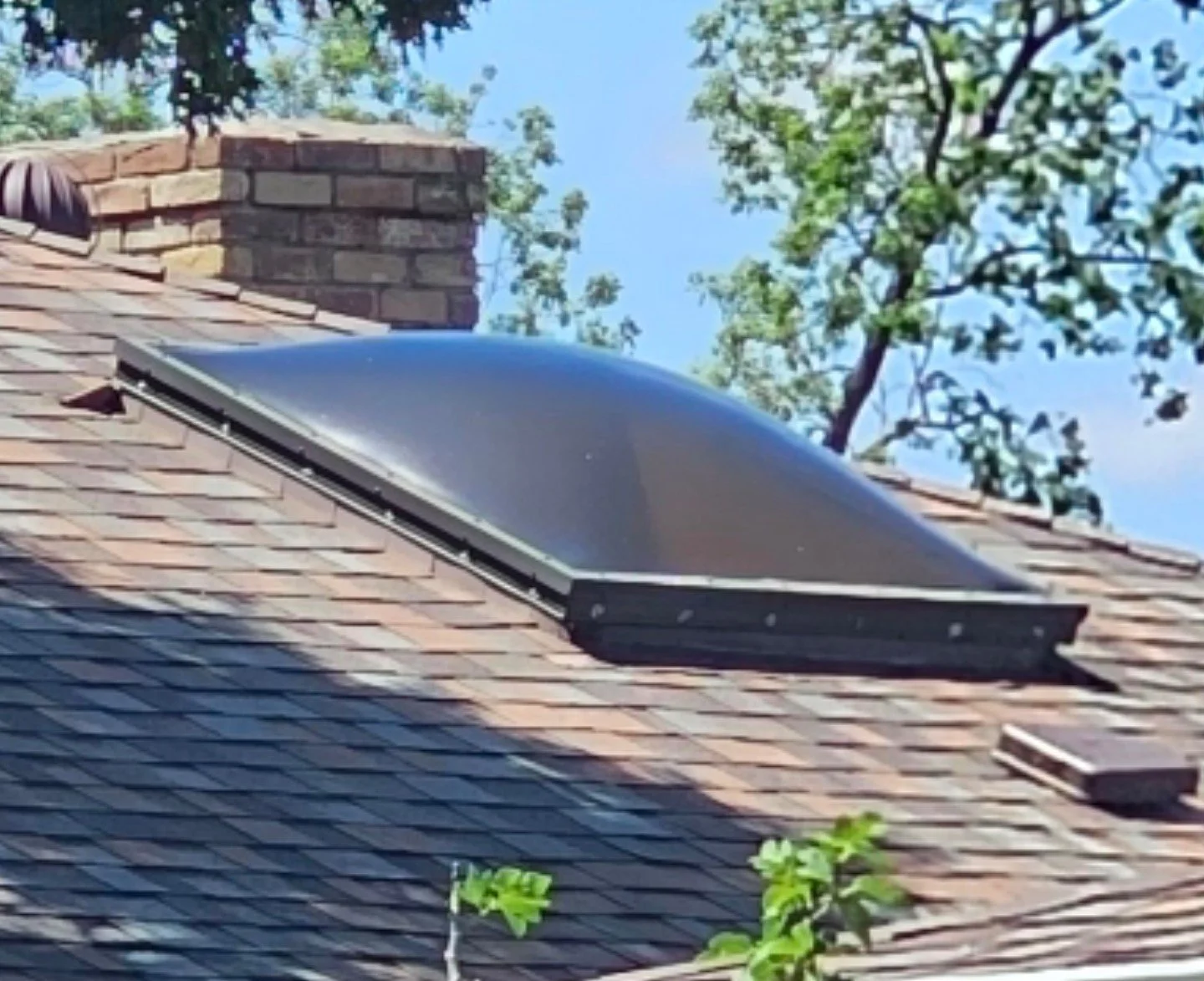 A solar attic fan installed on a residential shingled roof with trees and blue sky in the background.