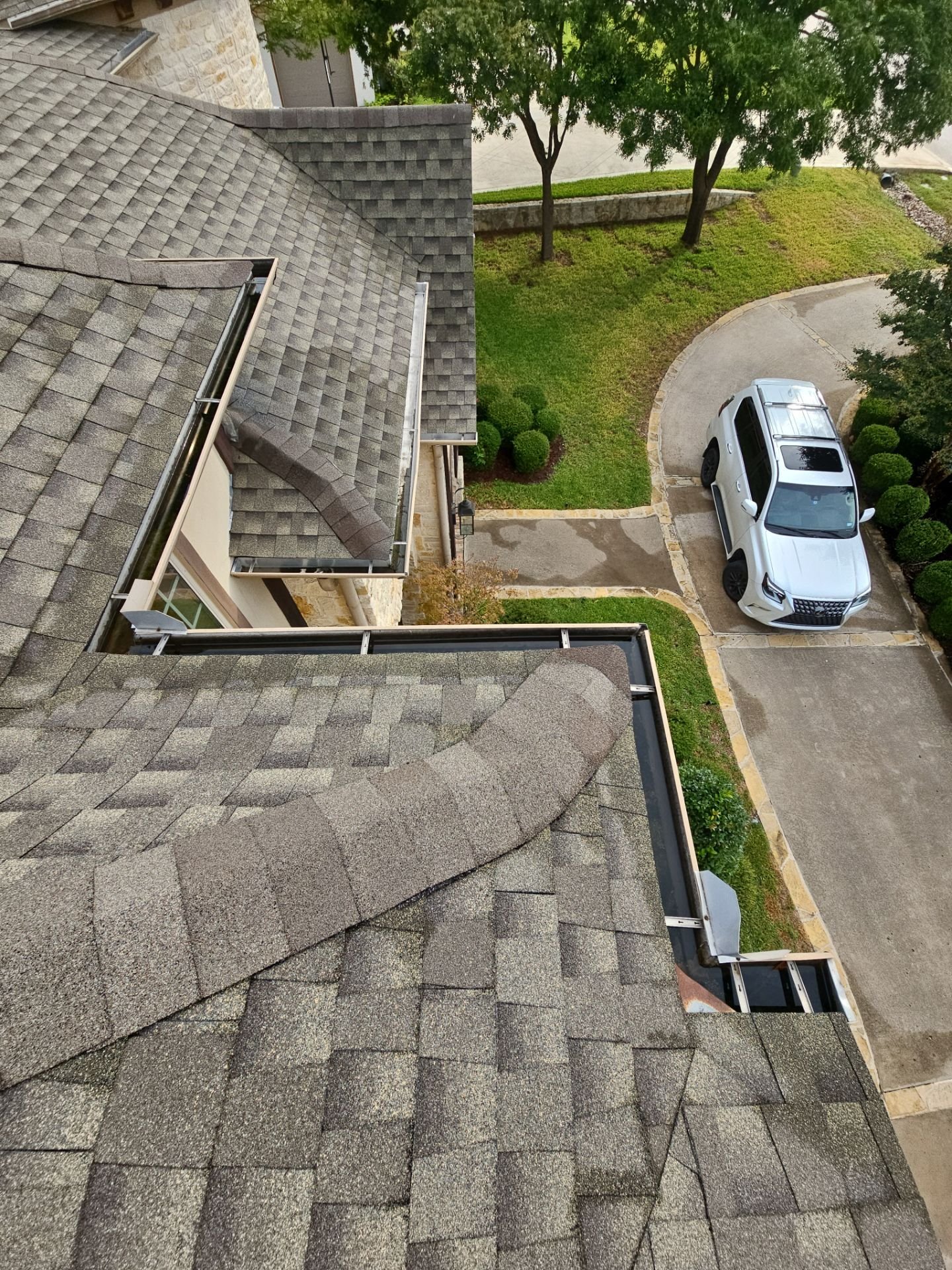 View from a high window showing a residential house roof with gray shingles, a well-maintained lawn, a curved driveway, and a white SUV parked on it, with some trees and shrubs nearby.