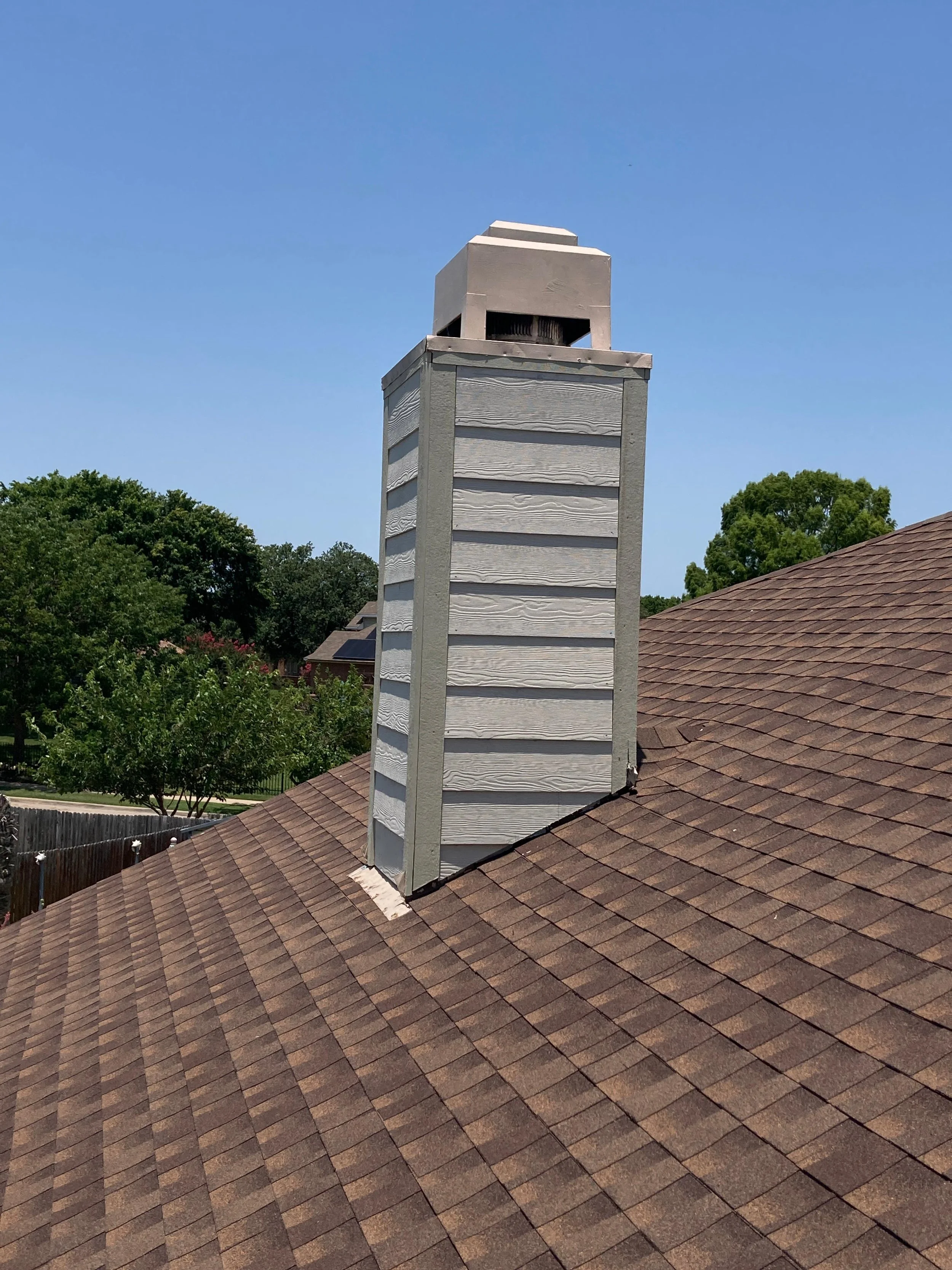 A house roof with a chimney and a chimney cap, under a clear blue sky with some trees in the background.