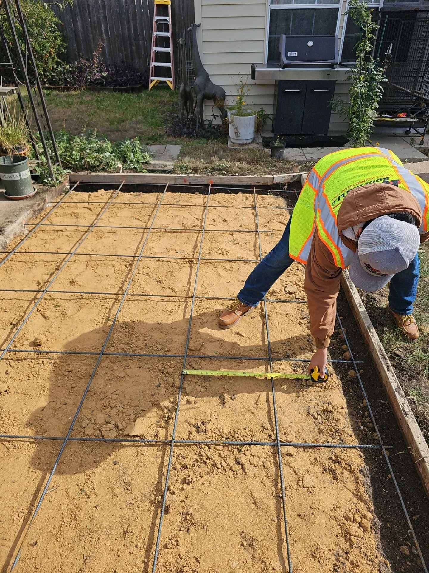 A person wearing a yellow safety vest, a gray cap, and work boots is measuring a wooden frame on a construction site in a backyard. There is a grid of rebar set into the ground, ready for concrete pouring.