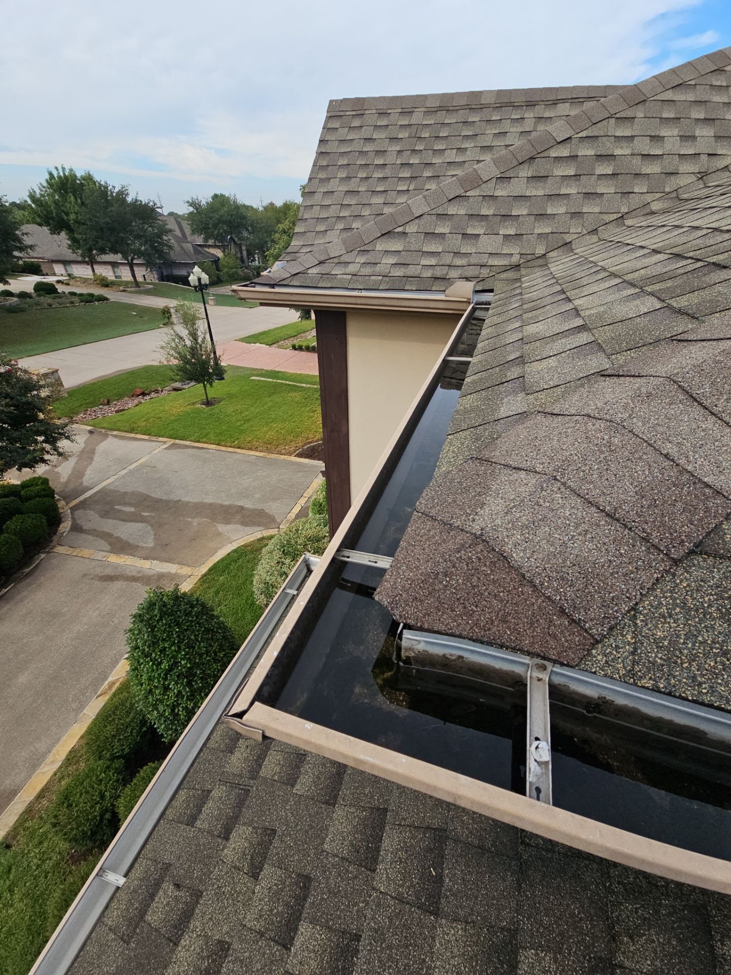 View of a residential roof with asphalt shingles, showing the gutters and surrounding neighborhood with houses, trees, and a parking lot.