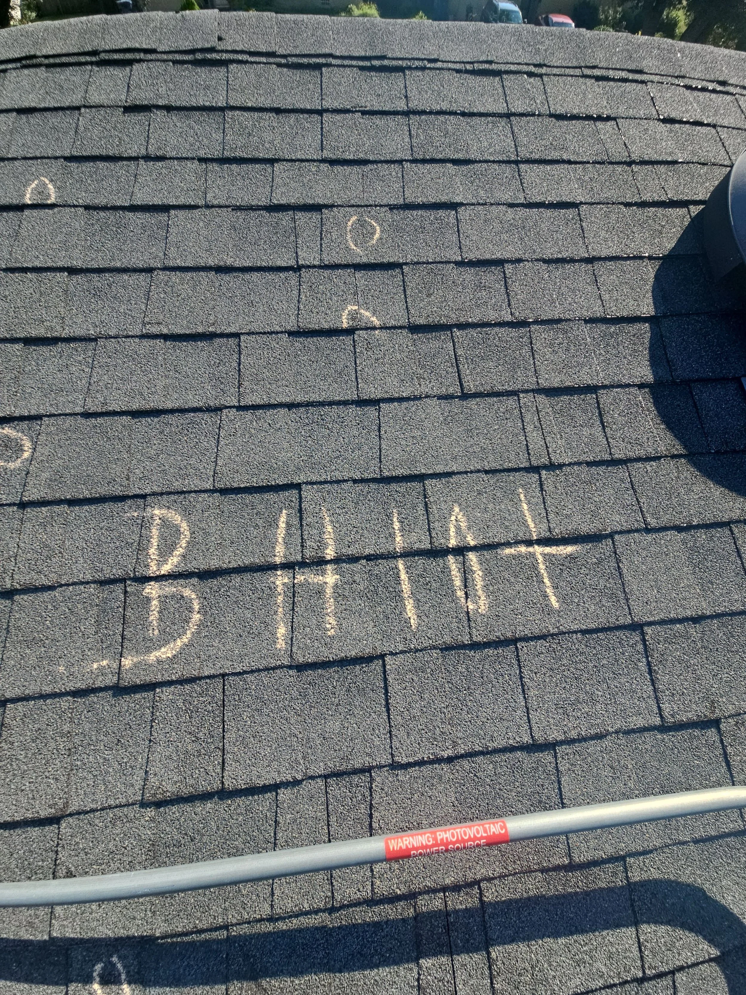 Roof shingles with chalk markings indicating areas for repair or inspection. A safety warning label about photovoltaic power source is visible on a pipe at the bottom.