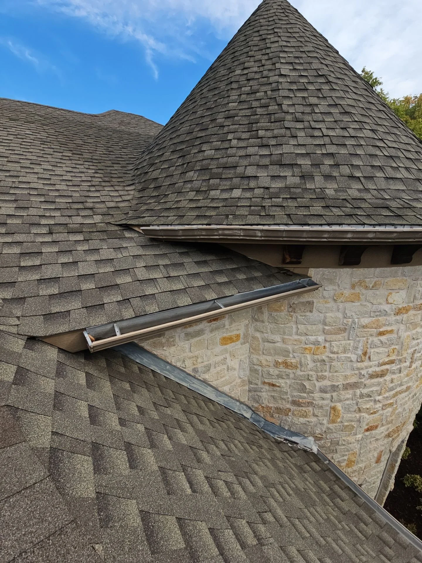 Close-up view of a house roof with asphalt shingles and a stone wall, against a partly cloudy sky.