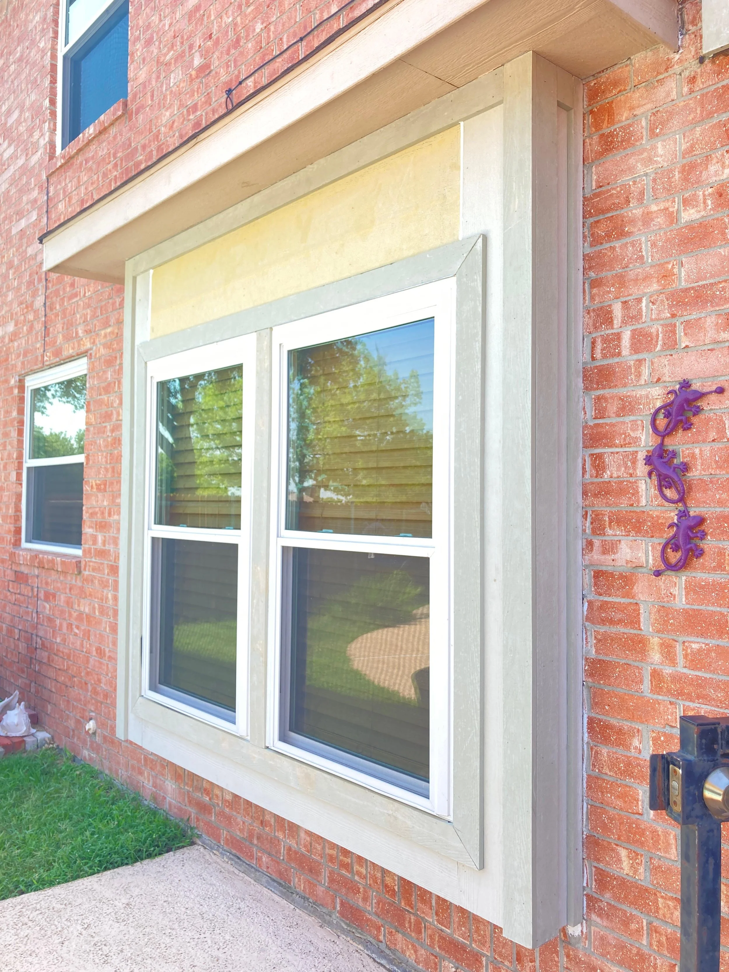 The exterior of a brick house with a large window with white framing, reflecting trees and the sky. Next to the window, purple decorative geckos are mounted on the brick wall. Part of a door frame and door handle are visible on the right side.