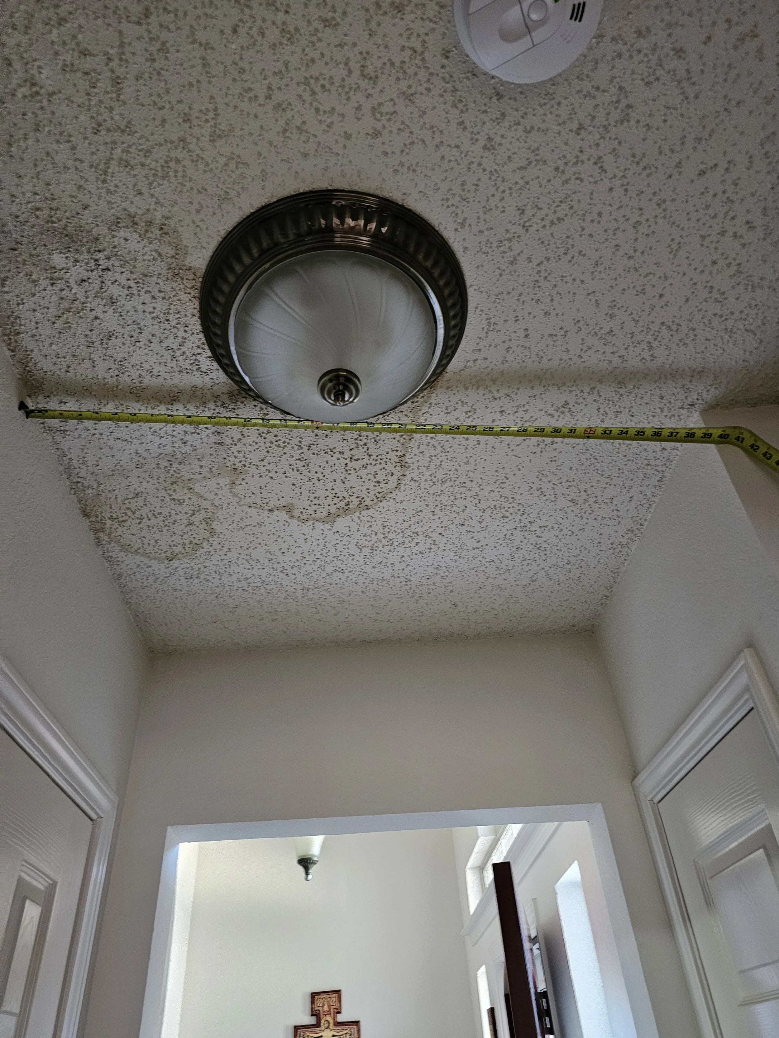A ceiling with textured surface and a smoke detector, a semi-flush light fixture, and a crown molding in a hallway leading to a well-lit room with a vaulted ceiling, a ceiling fan, and decorative art on the wall.