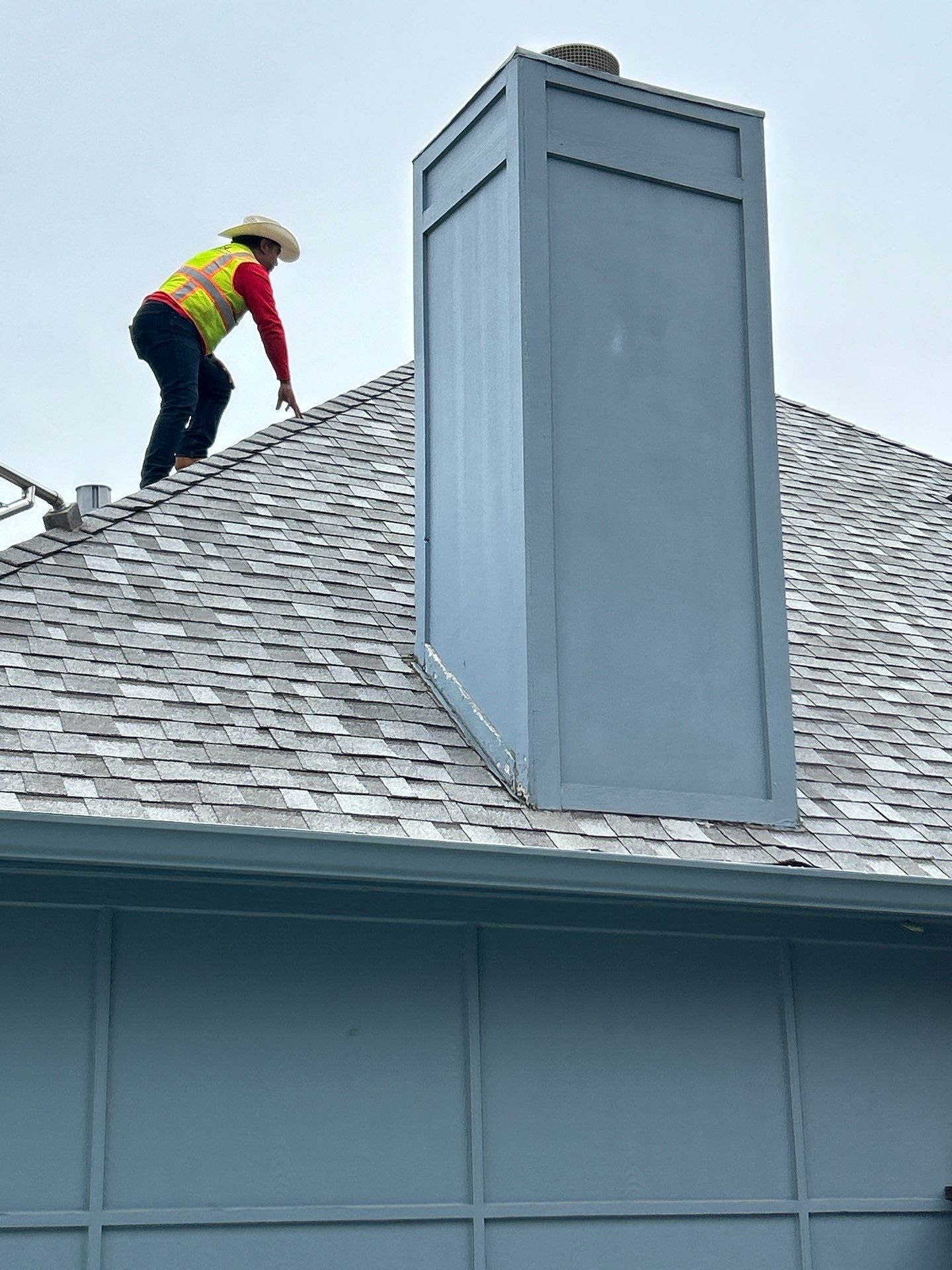 A person wearing a cowboy hat, red shirt, reflective safety vest, and dark pants crouches on a roof, near a large blue chimney.