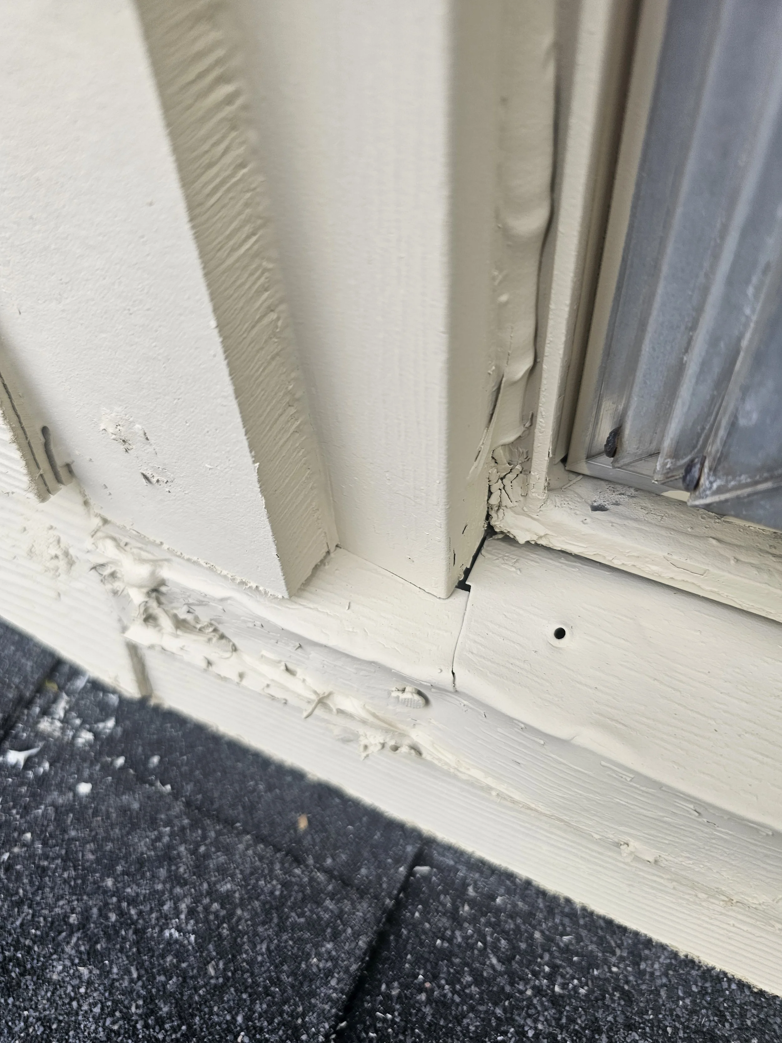 Close-up of a white door frame with peeling paint and a damaged corner, showing the black asphalt ground outside.