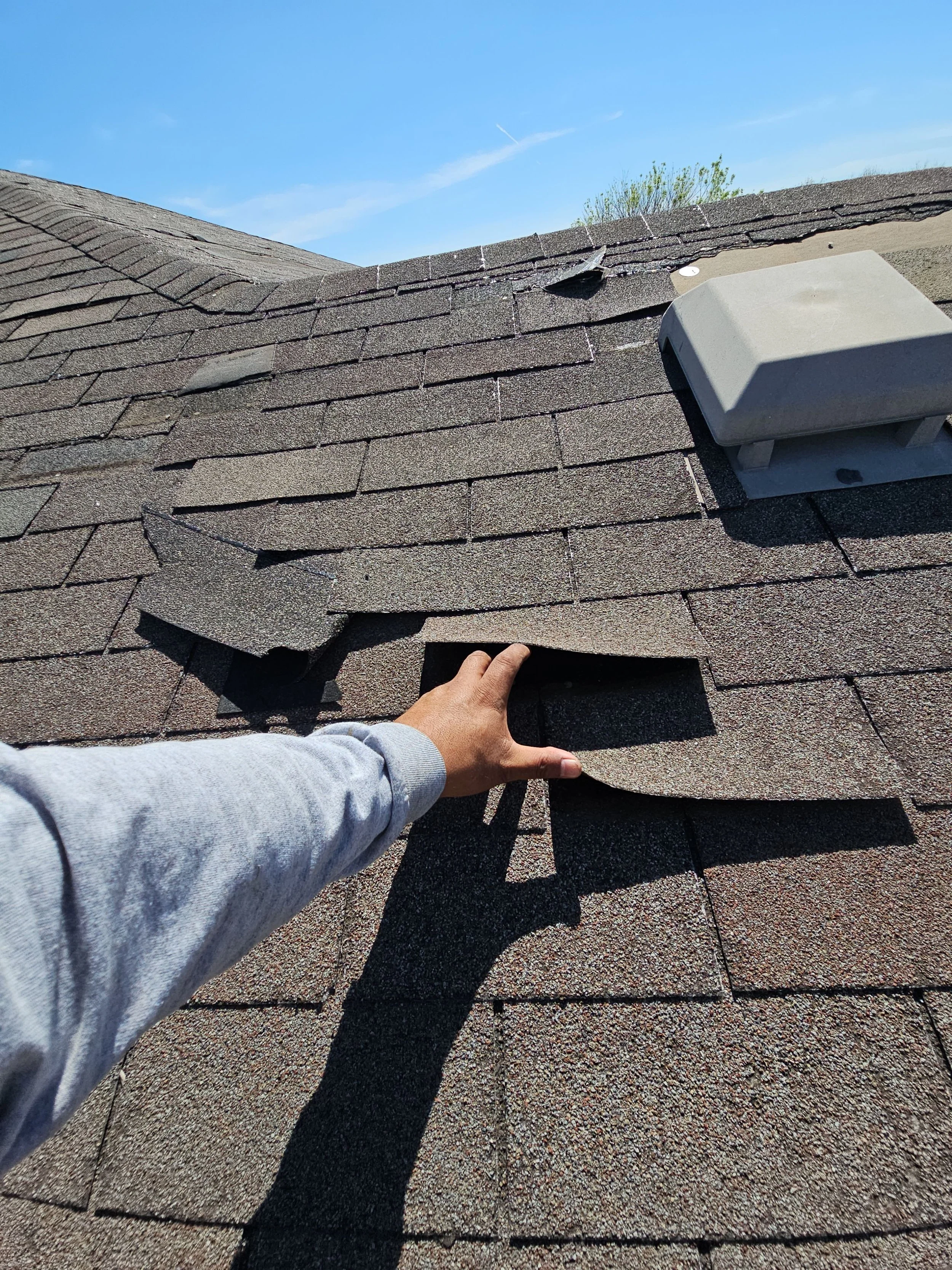 Person inspecting a shingle roof under blue sky with some trees in the background.