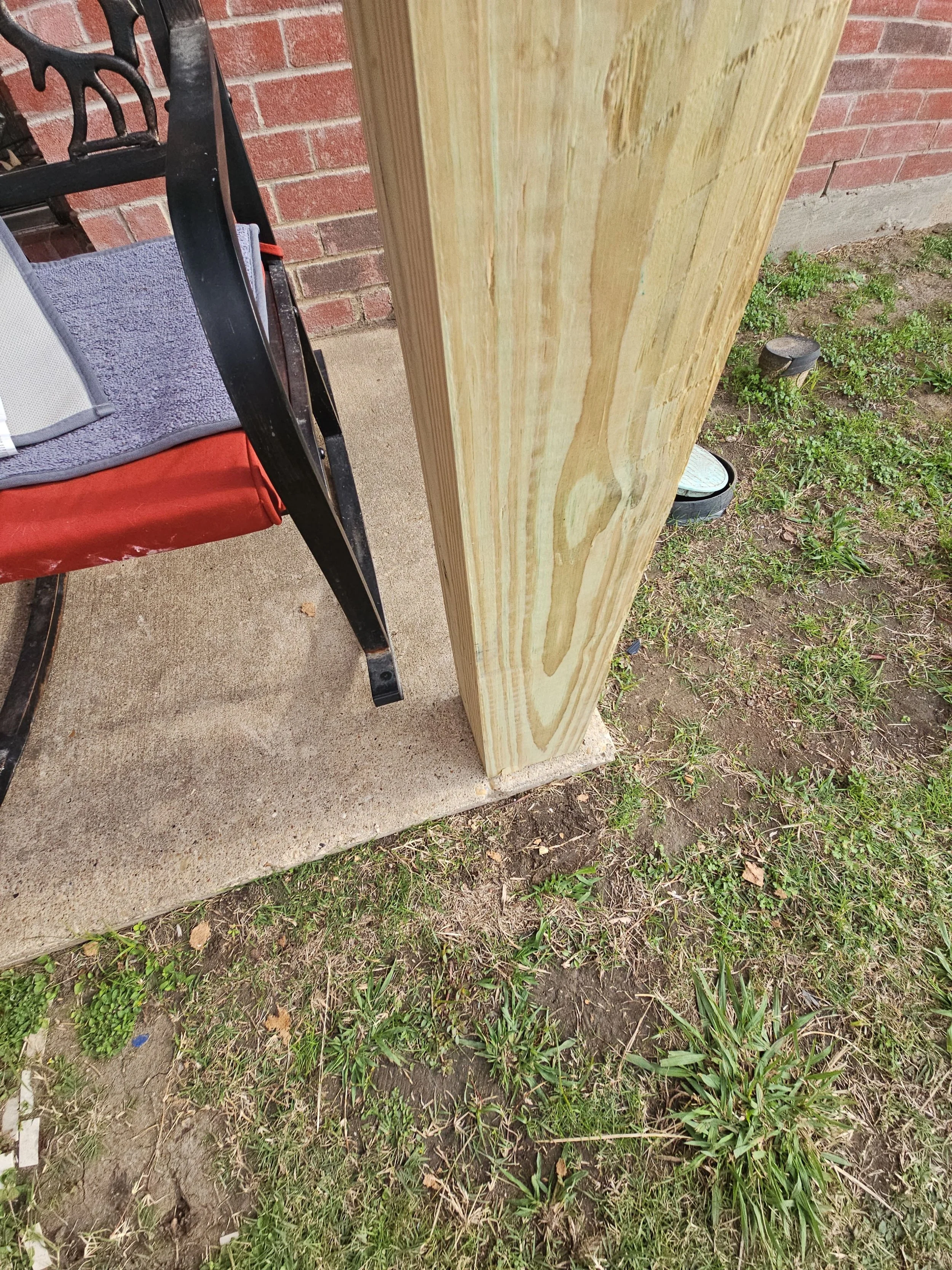 A close-up view of a partially constructed wooden post installed on concrete, with a lawn and a brick wall in the background, and an outdoor light fixture on the ground nearby.