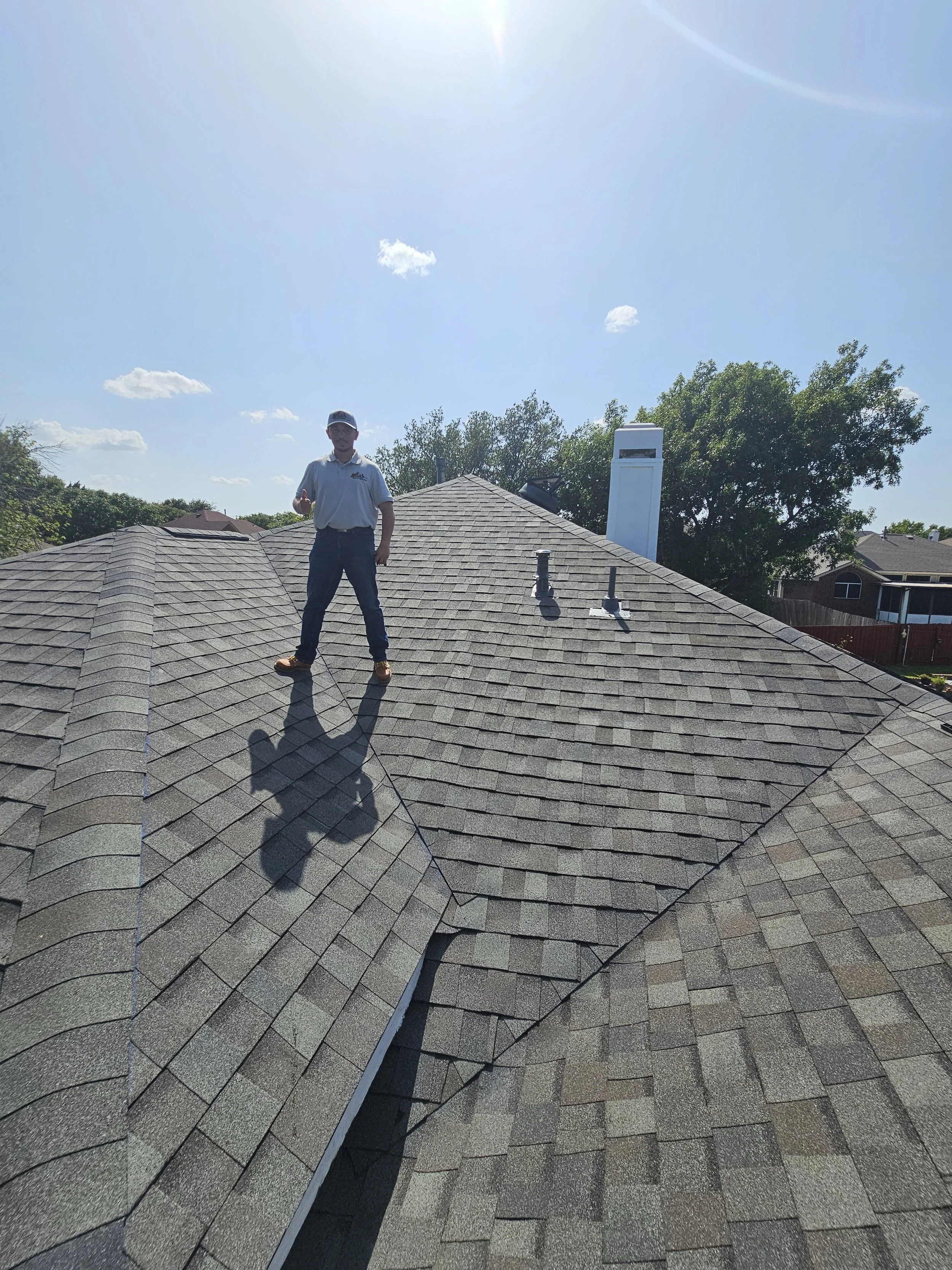 A man standing on a roof with gray shingles, giving a thumbs-up gesture, under a bright sky with a few scattered clouds and sunlight.