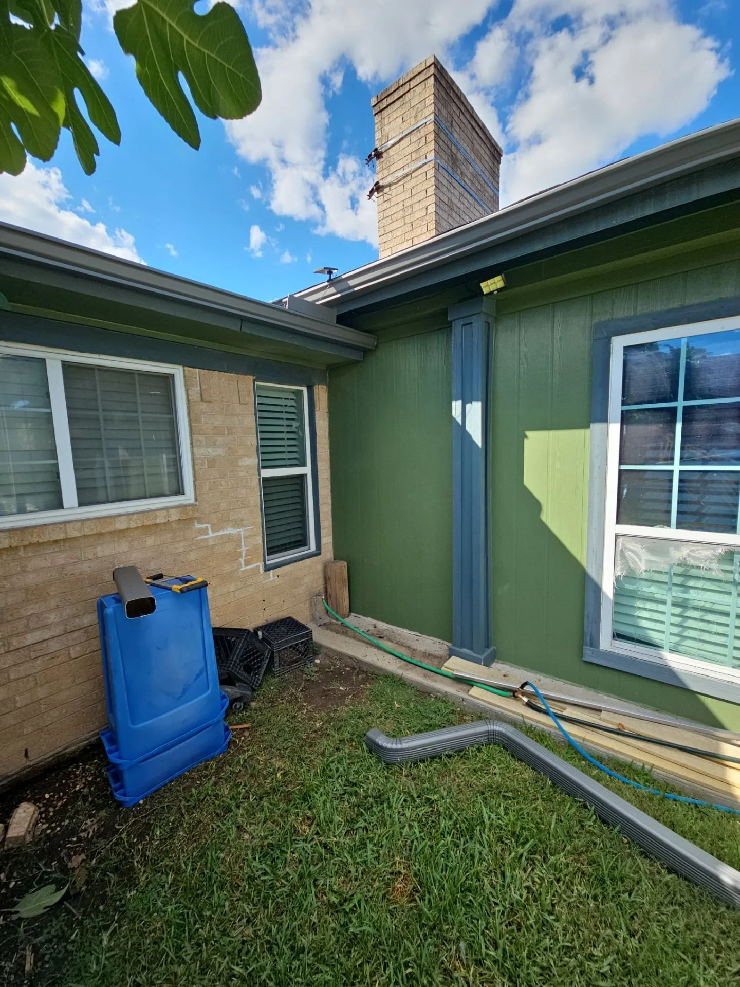 Backyard with green house wall, window, and construction materials, including a blue trash bin, black crates, and pipes, on a patchy grass yard with a brick house wall and a chimney in the background under a blue sky with clouds.