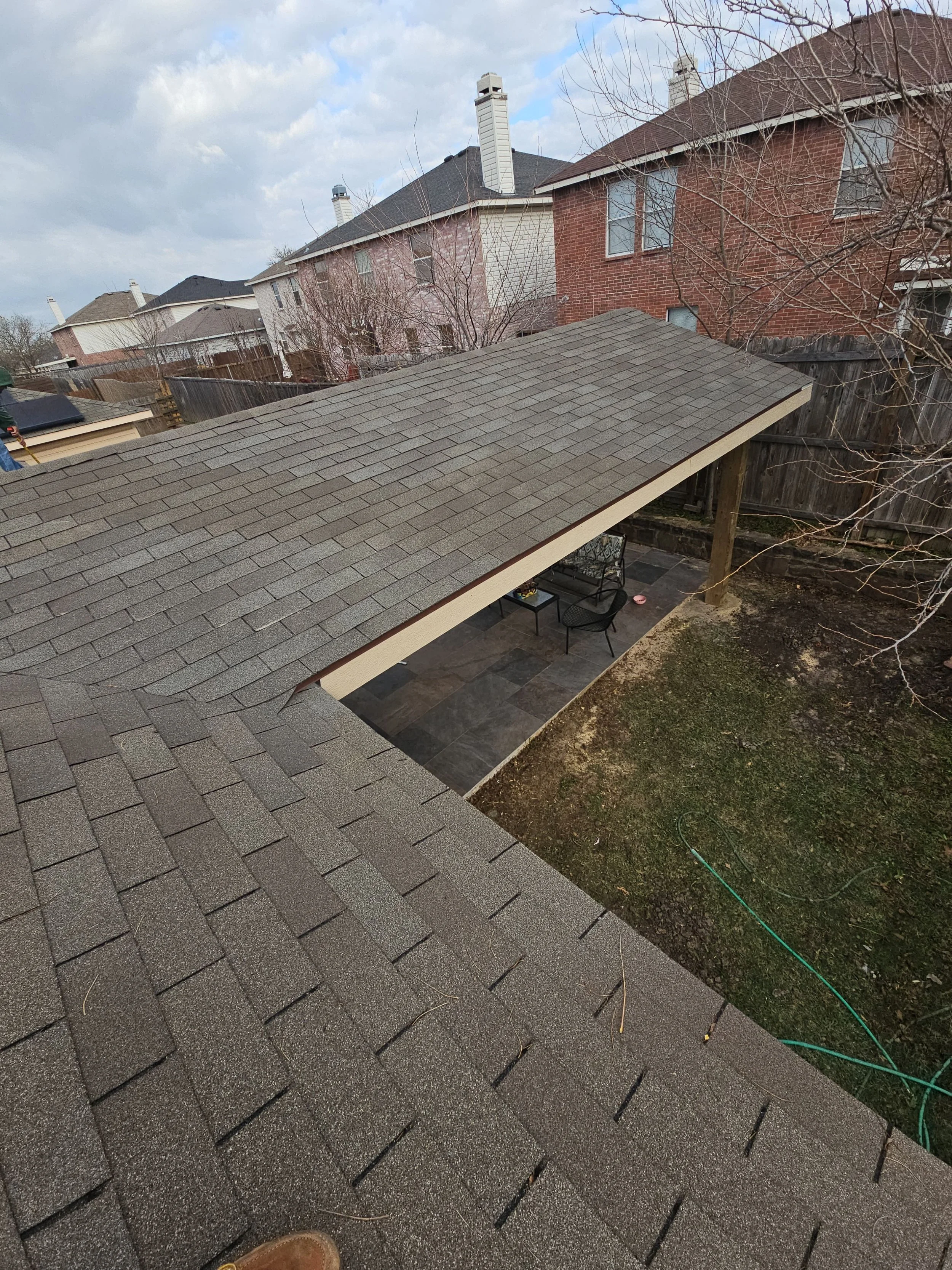 View of rooftops of houses and a backyard patio with chairs. Roofs have asphalt shingles; some houses have brick walls and others have siding. Bare trees and a cloudy sky are visible.