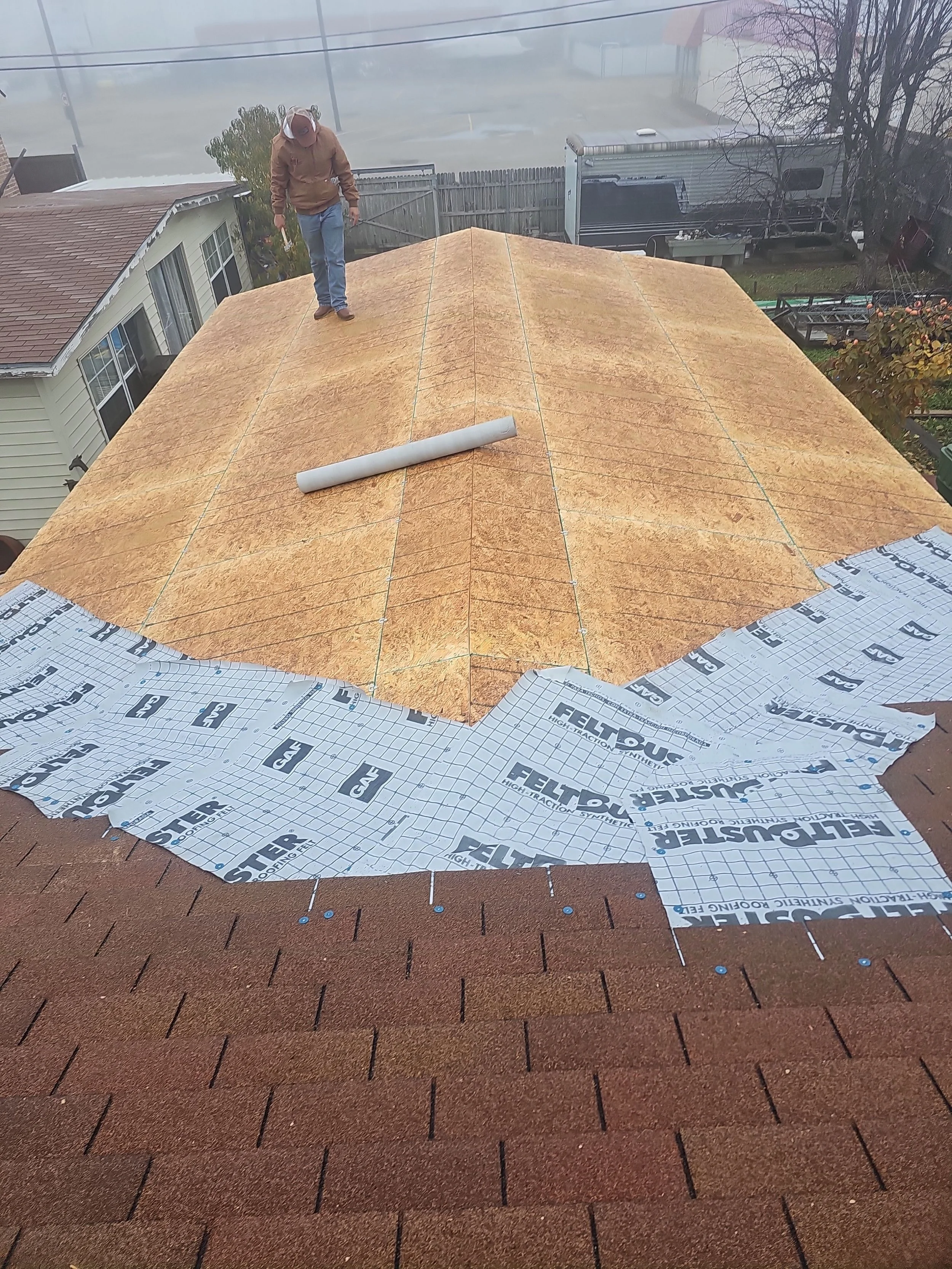 A person working on installing a new roof, with sheets of roofing underlayment and asphalt shingles, on a residential house's roof, on a foggy day.