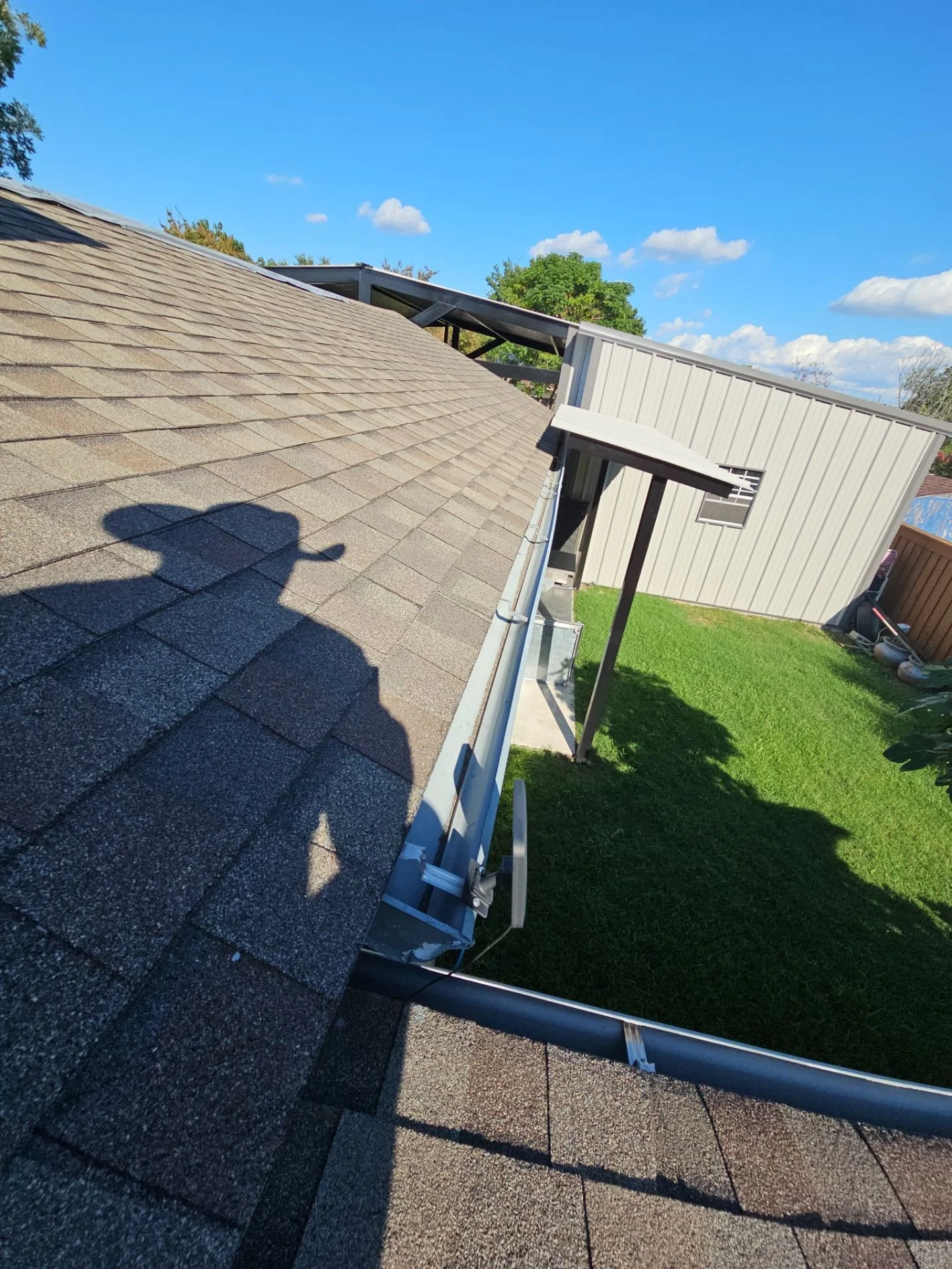 Photo of a house roof with new shingles and solar panel installation, green lawn, blue sky with clouds, and part of a backyard with garden tools.
