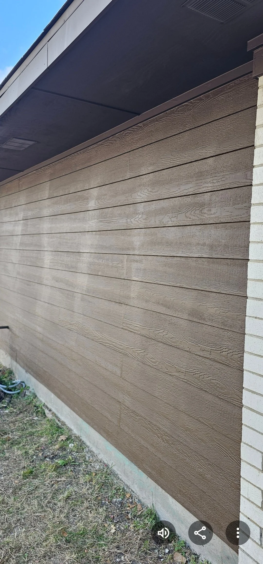 Side view of a house exterior showing beige horizontal siding, a small patch of grass and dirt, and part of a white brick wall.