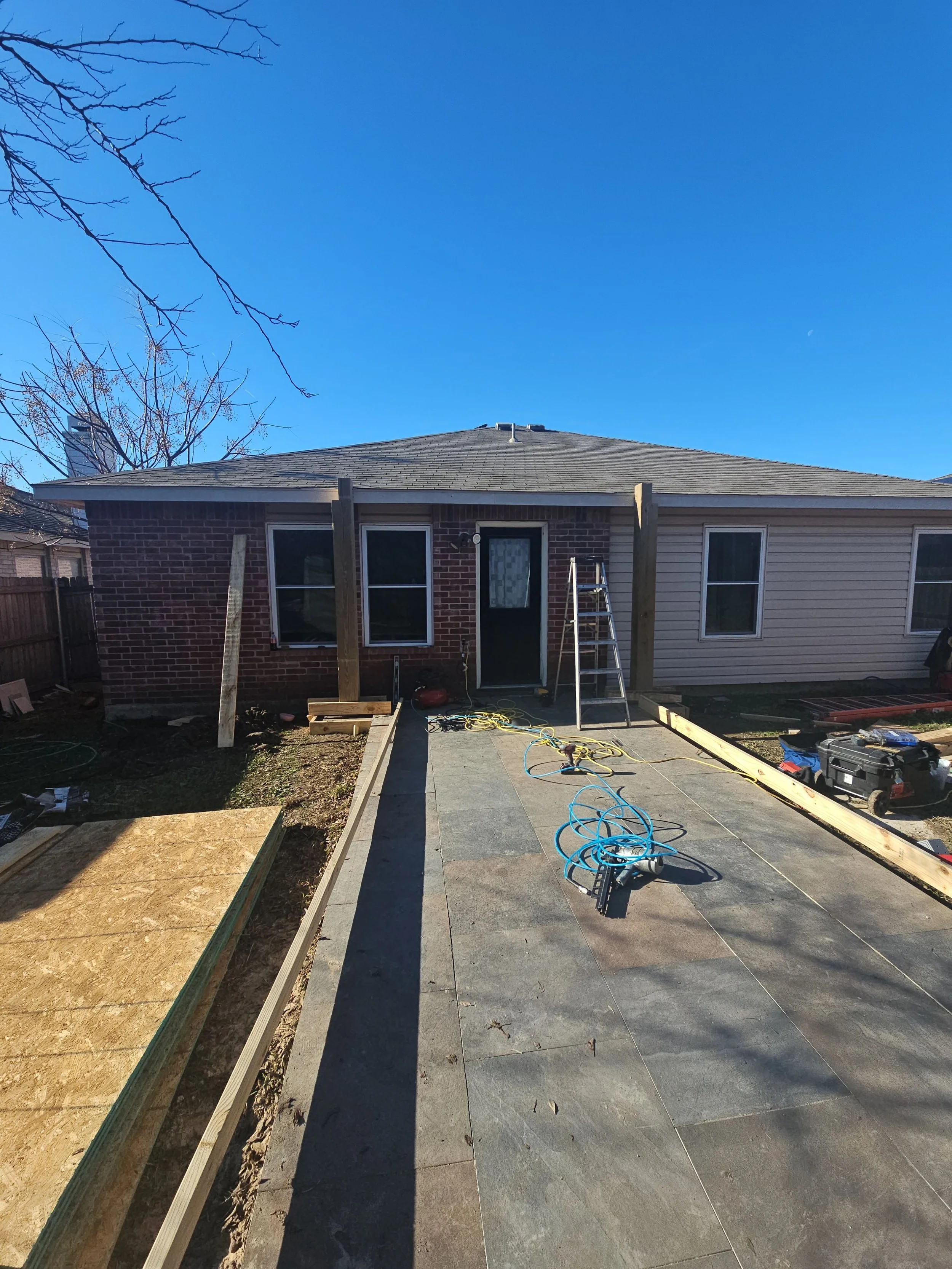 Backyard with house under construction, with wooden framing, construction tools, ladder, and partially completed walkway.