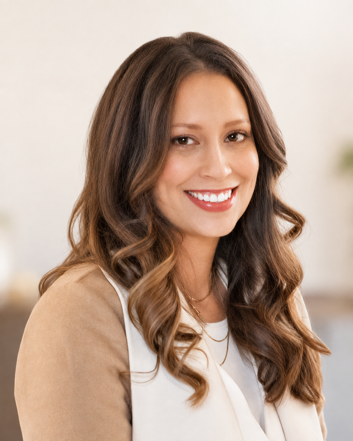 A woman with long, wavy brown hair and a bright smile, wearing a beige blazer over a white top, in a softly lit indoor setting.
