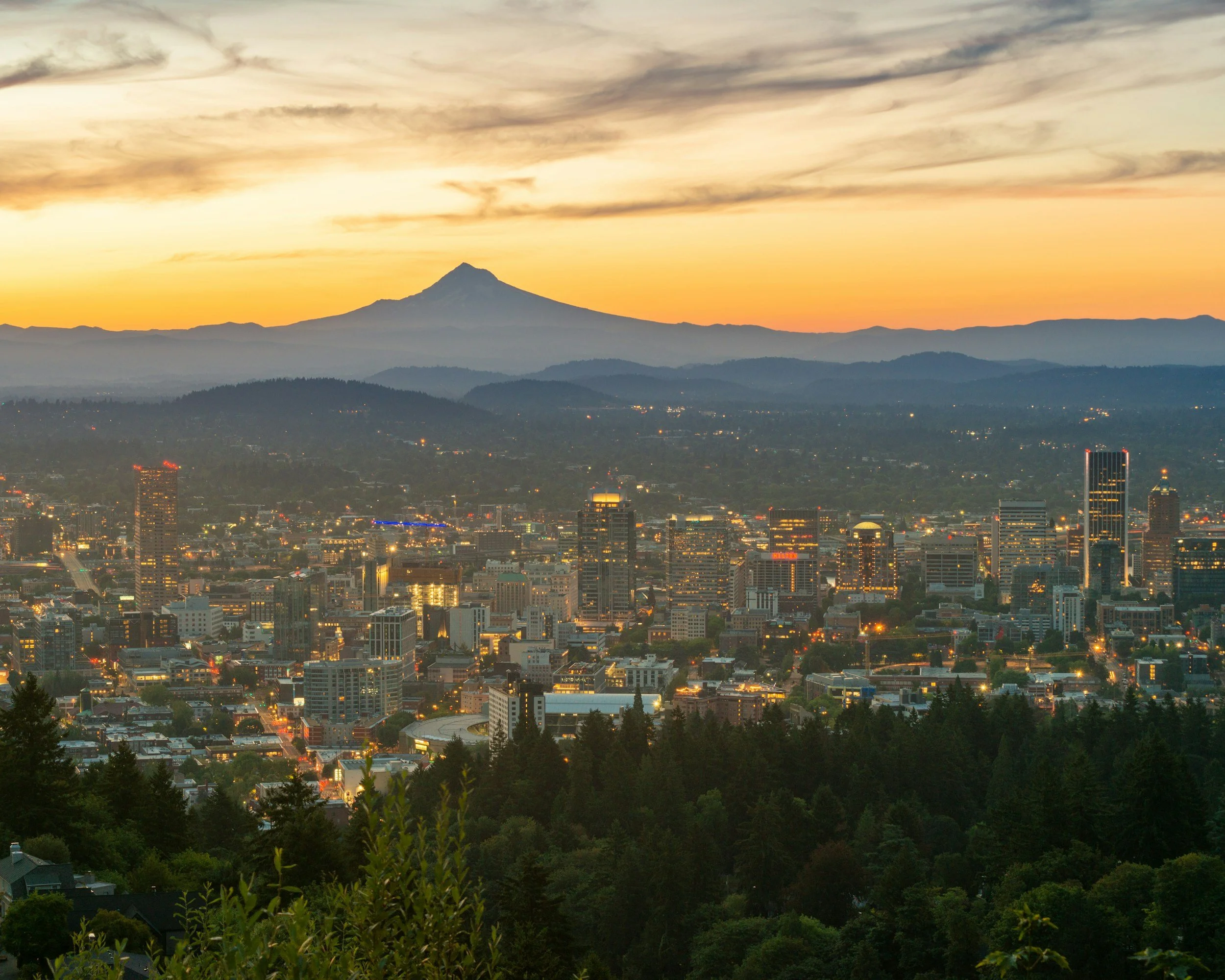 Portland cityscape at sunset with the silhouette of Mount Hood in the background, clouds in the sky, and city lights beginning to turn on across the city.