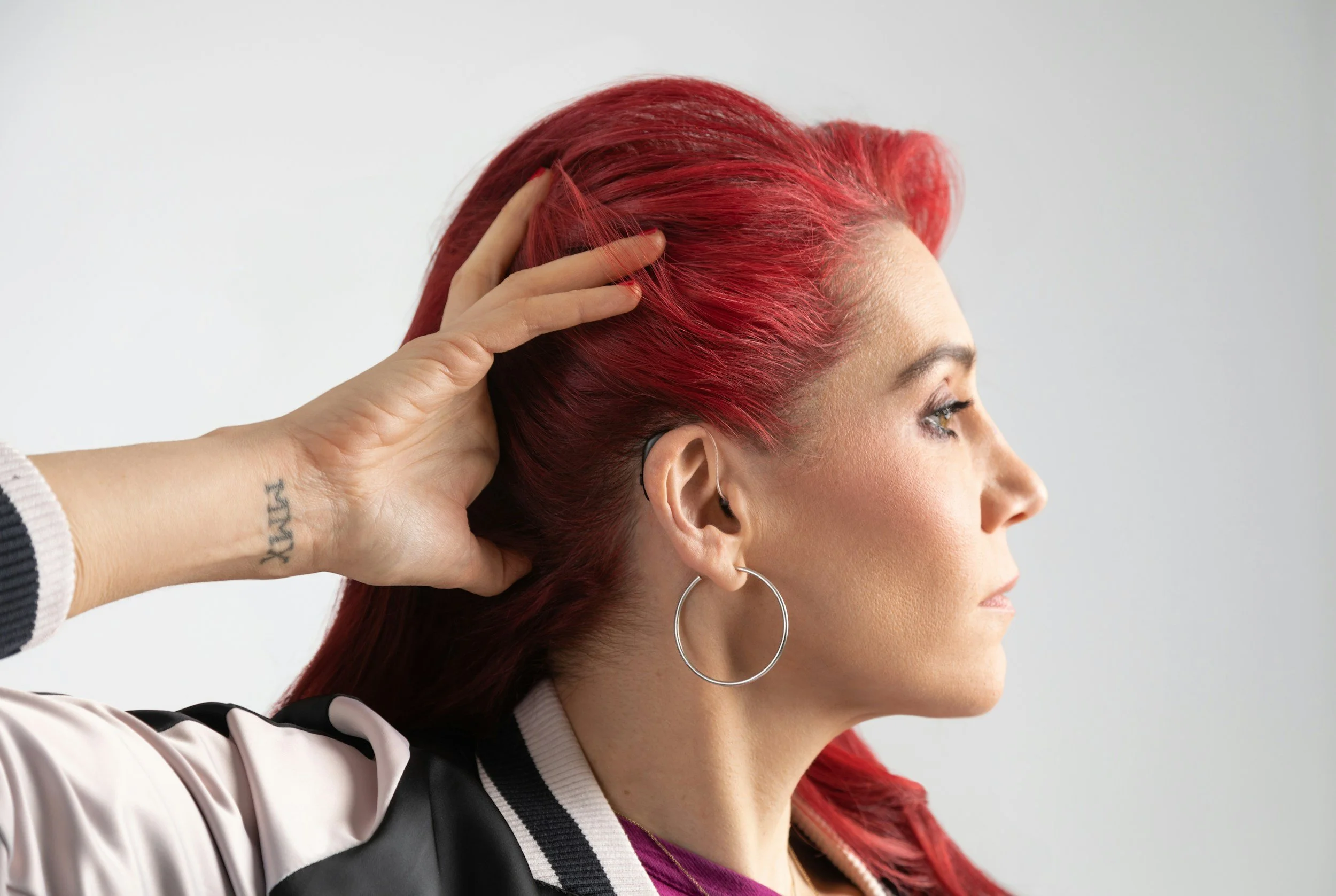 A woman with red hair, wearing hearing aids and large hoop earrings, with her head turned to the side. A hand with a tattoo on the wrist is holding back her hair revealing her hearing aid.